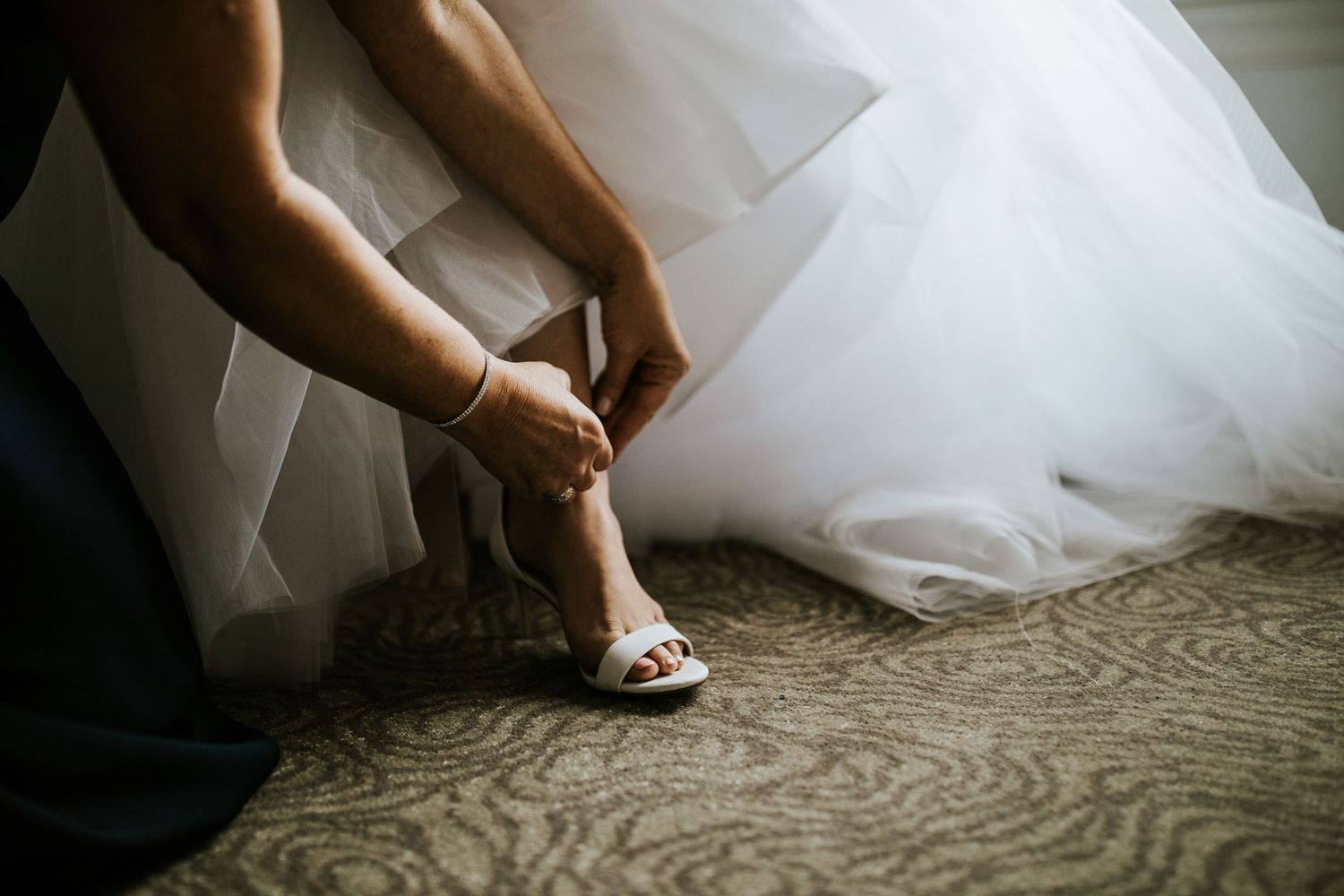 Close-up of delicate bridal shoes being fastened with tulle dress fabric in the background.