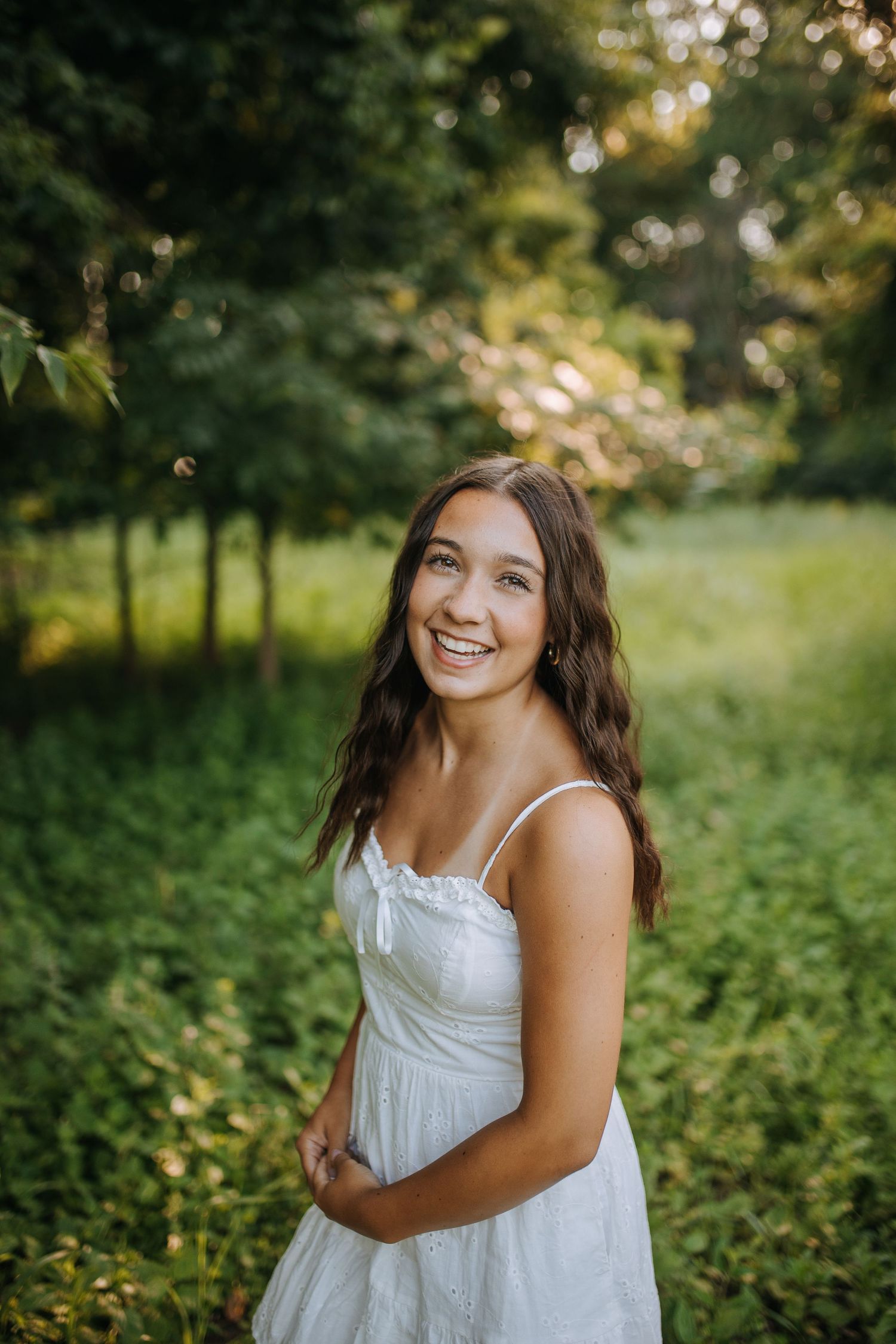 Person in white sundress poses in natural outdoor setting with green foliage.