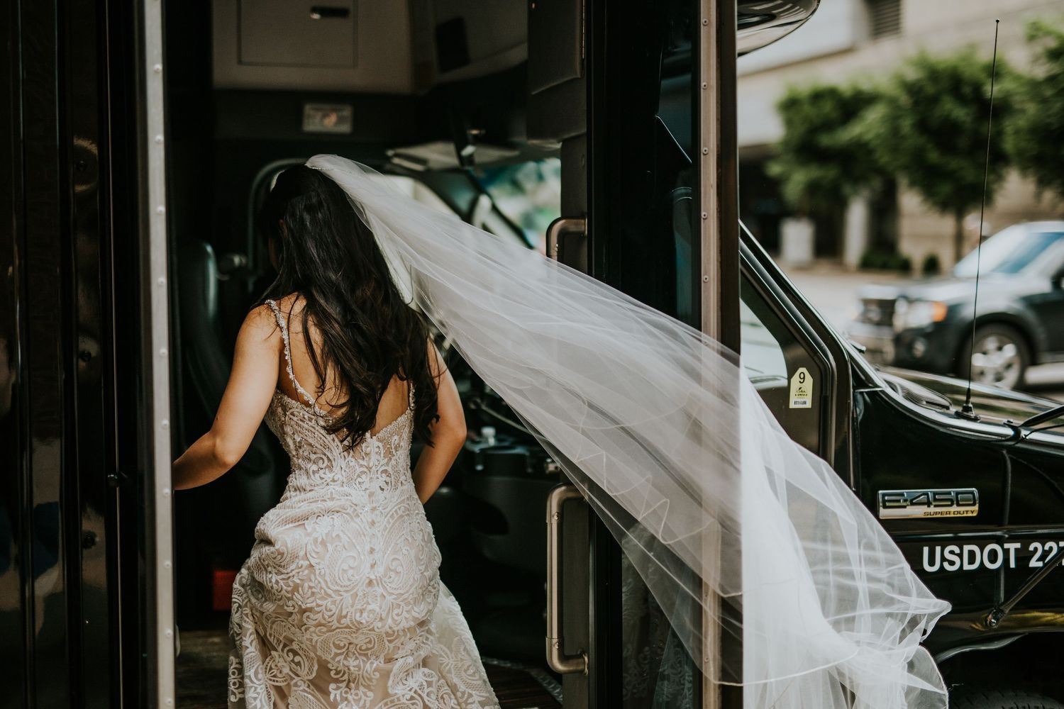 Long flowing wedding veil catches the breeze as bride in lace dress enters a vehicle.