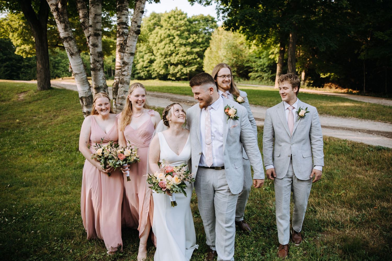 A bridal party in pink and gray attire walks together beneath birch trees.