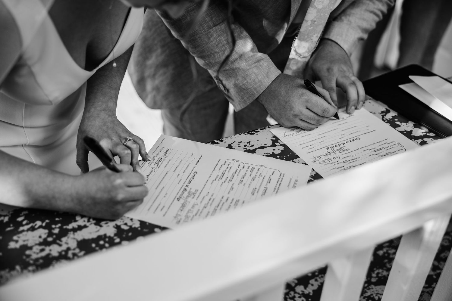 Black and white photo of hands signing wedding documents on a patterned tablecloth.