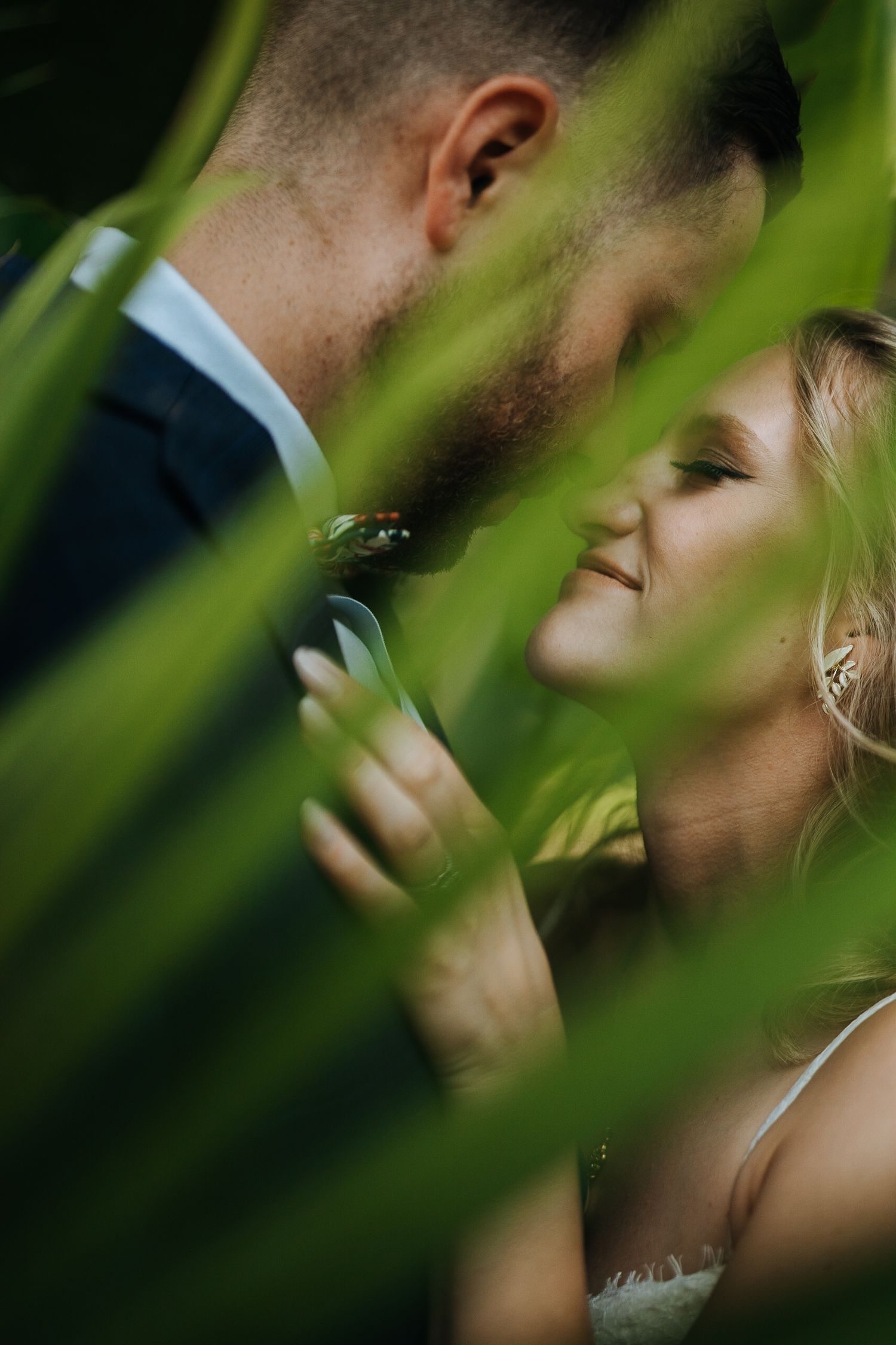 Romantic wedding moment captured through green tropical foliage in a greenhouse setting.