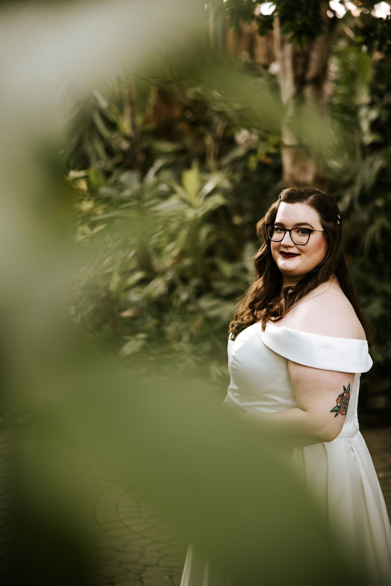 Bride in white off-shoulder dress poses among lush greenery.