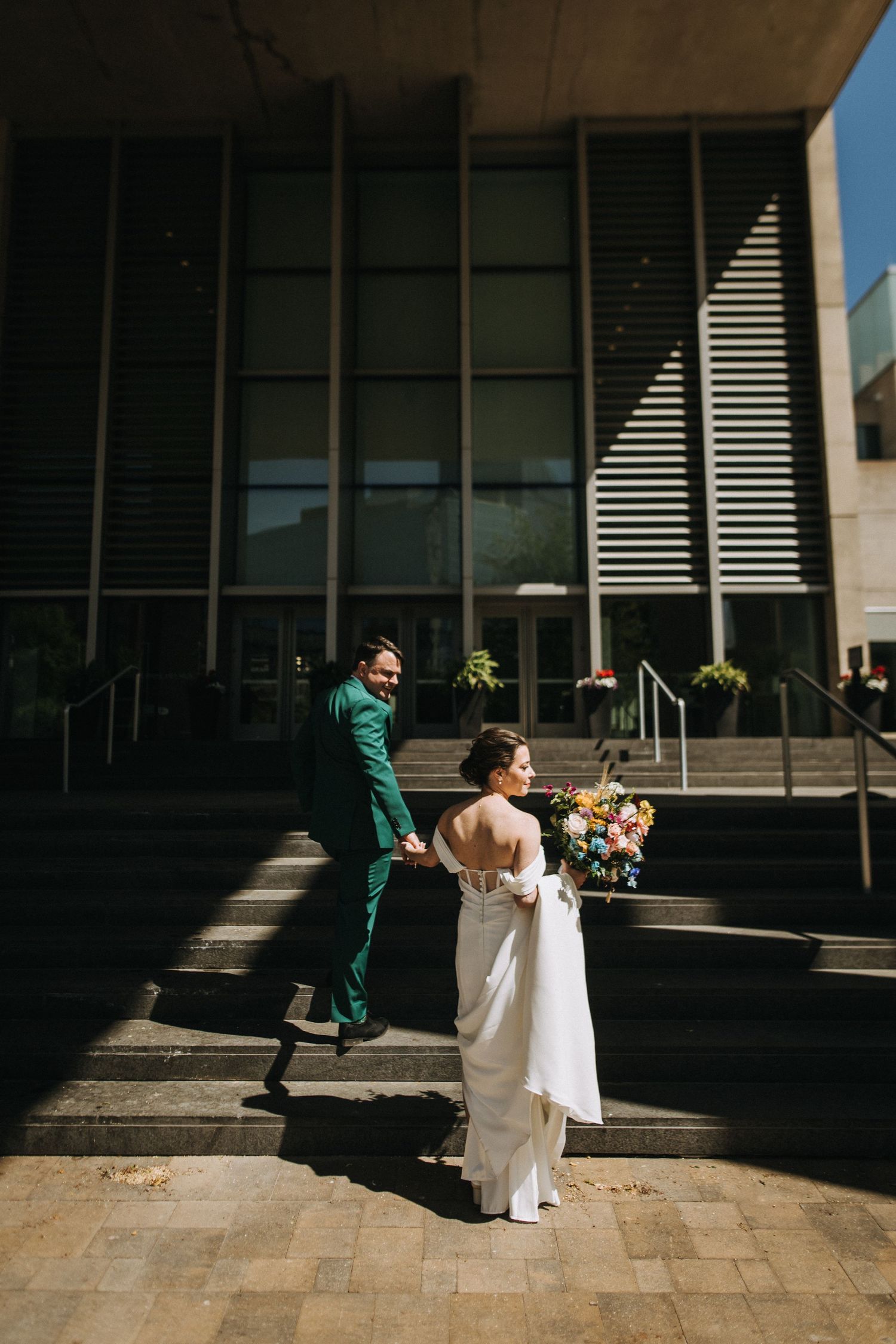 Bride in white gown with bouquet and groom in green suit walk up steps outside modern building on wedding day.