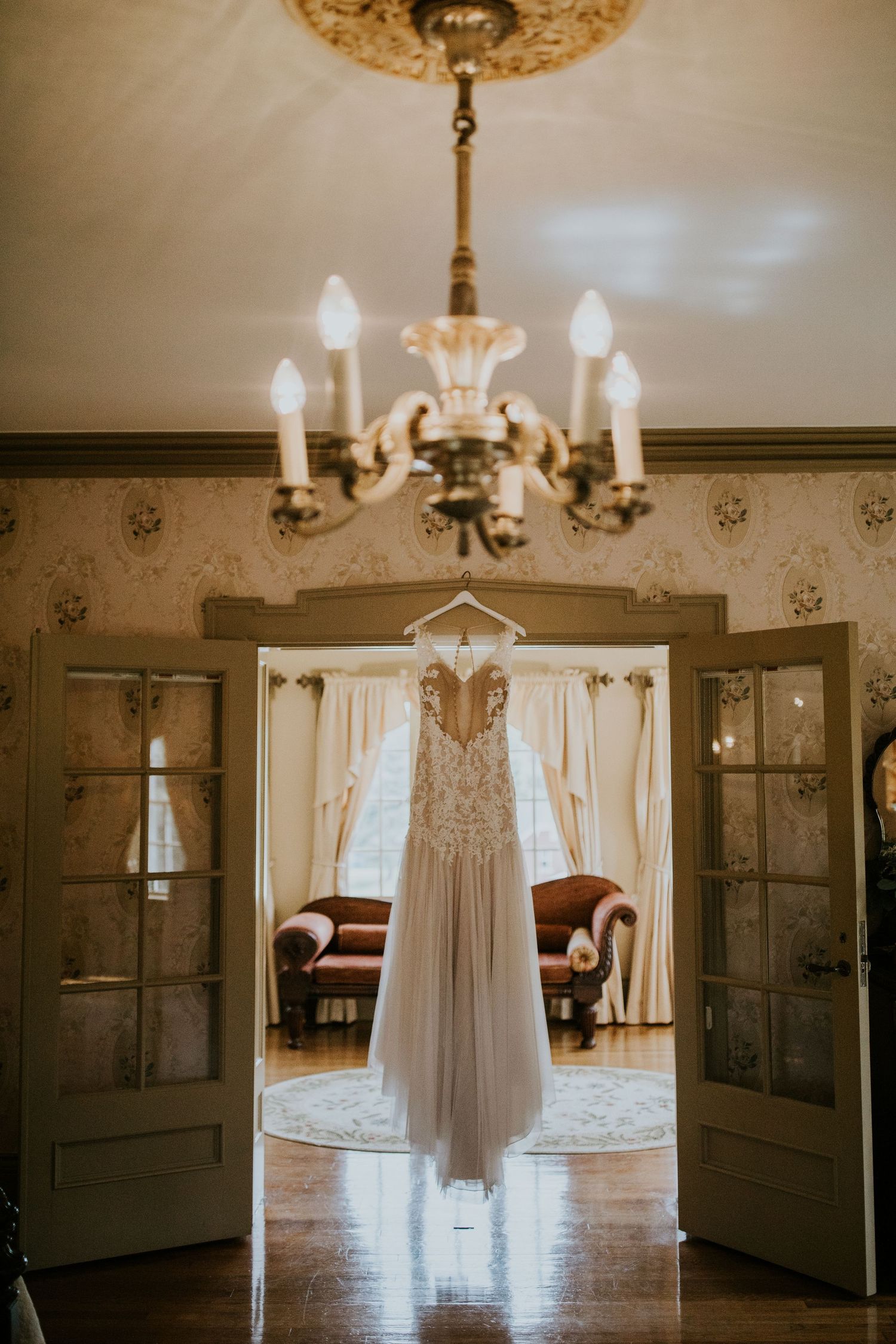 Elegant white wedding dress hangs from chandelier in vintage room with natural light streaming through windows.