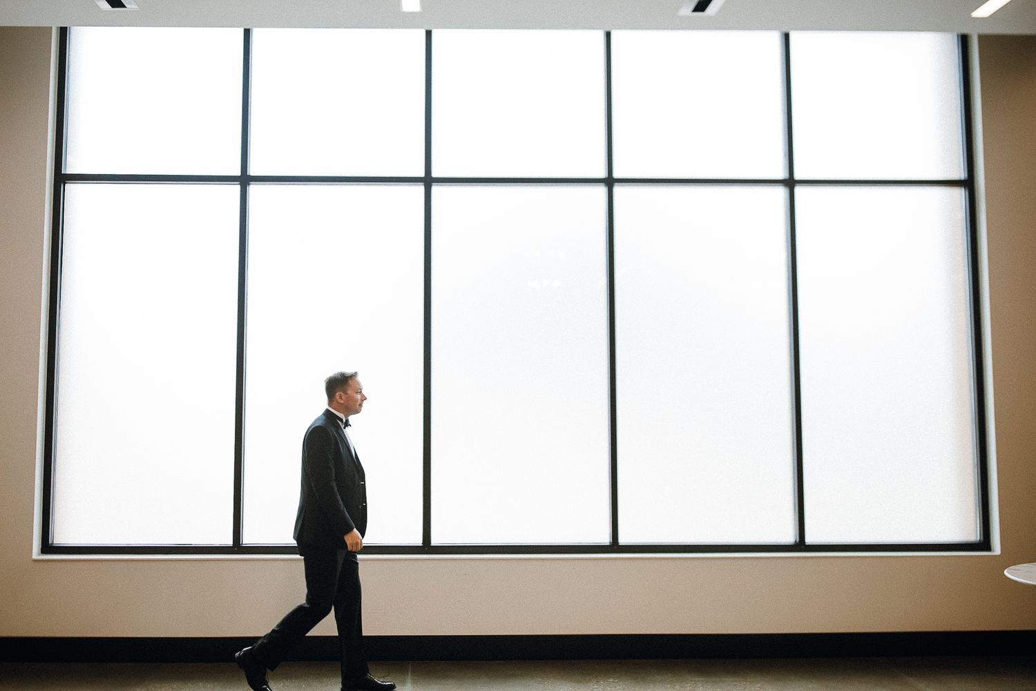 Silhouette of a person in formal attire walking past large modern windows with bright natural light.