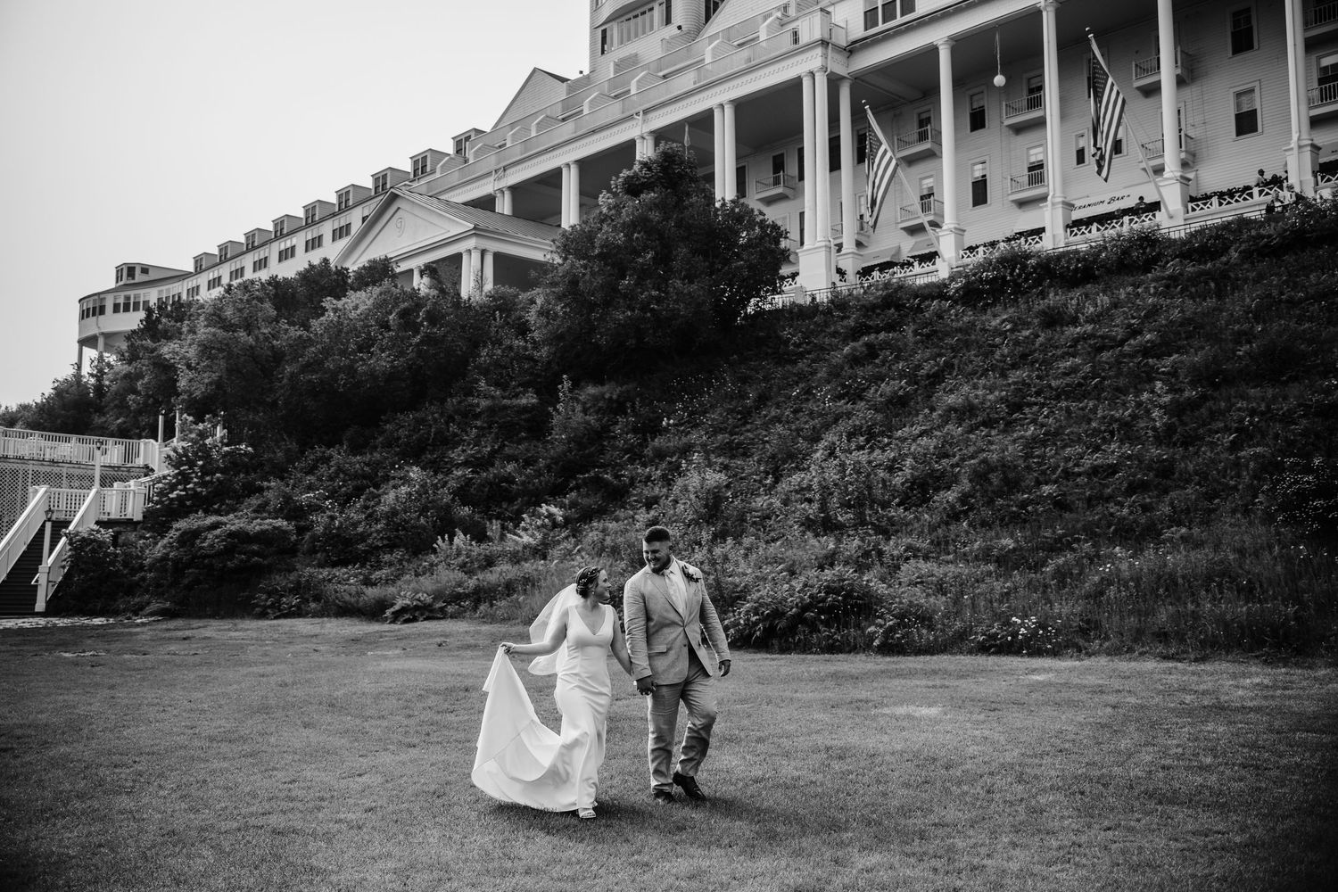 Black and white photo of couple walking on lawn in front of grand historic hotel.
