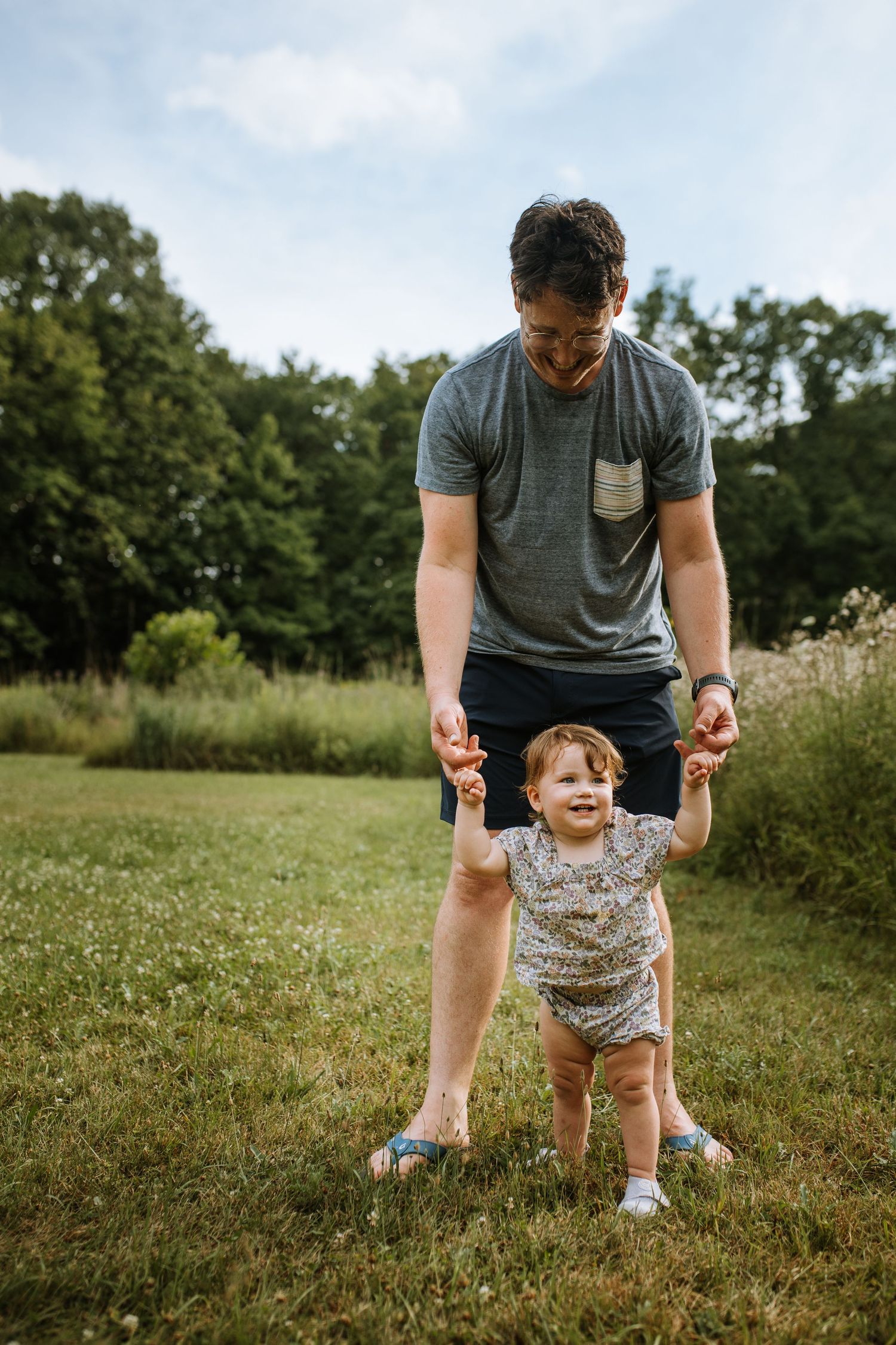 Parent helps small child take first steps on grassy field during summer day.