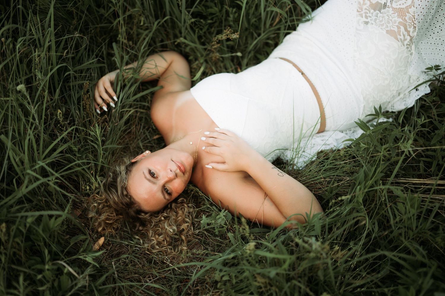 Person in white dress laying in tall grass during an outdoor photo session.
