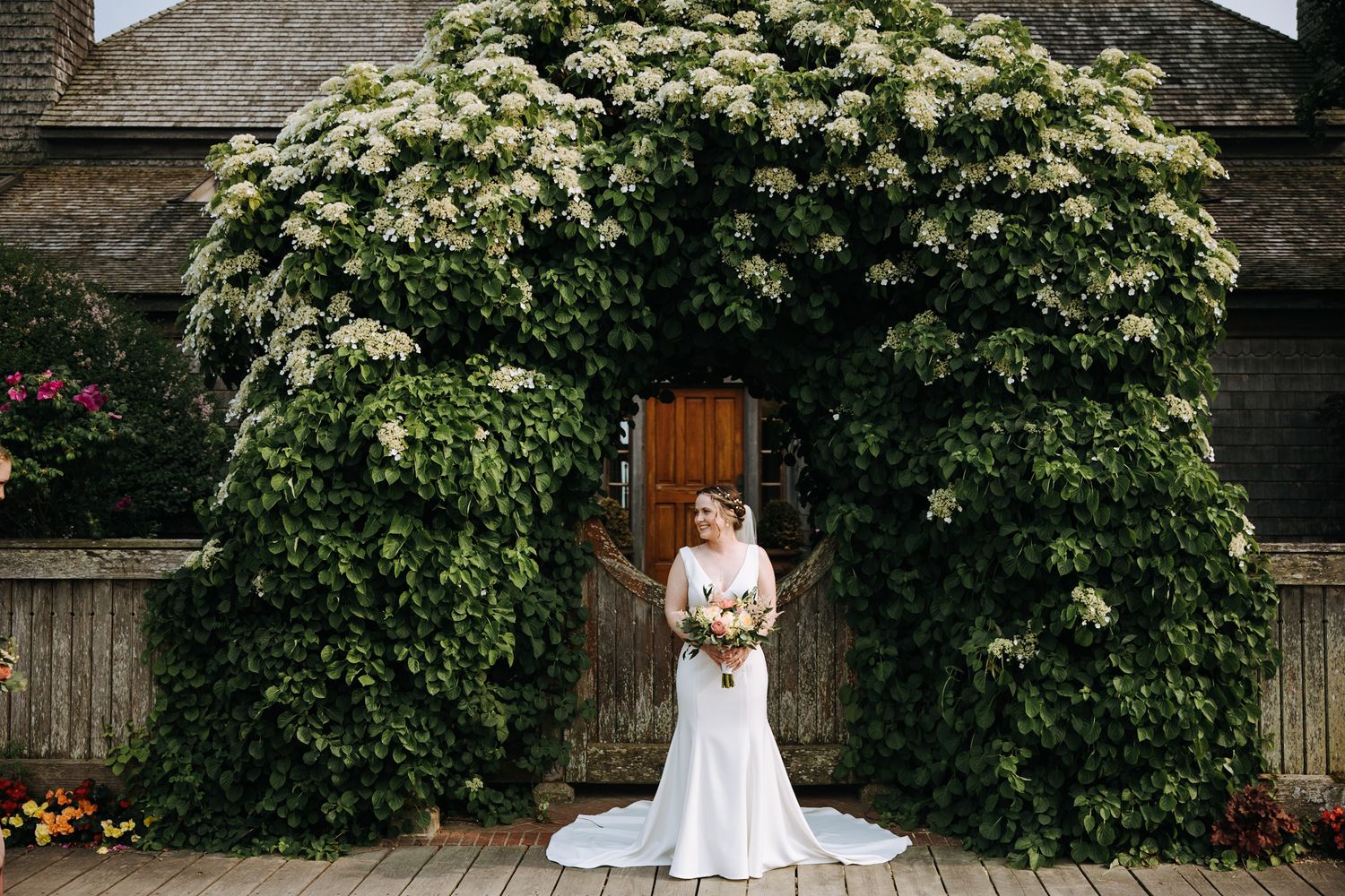 A bride stands beneath a flowering archway entrance with greenery and white blooms.