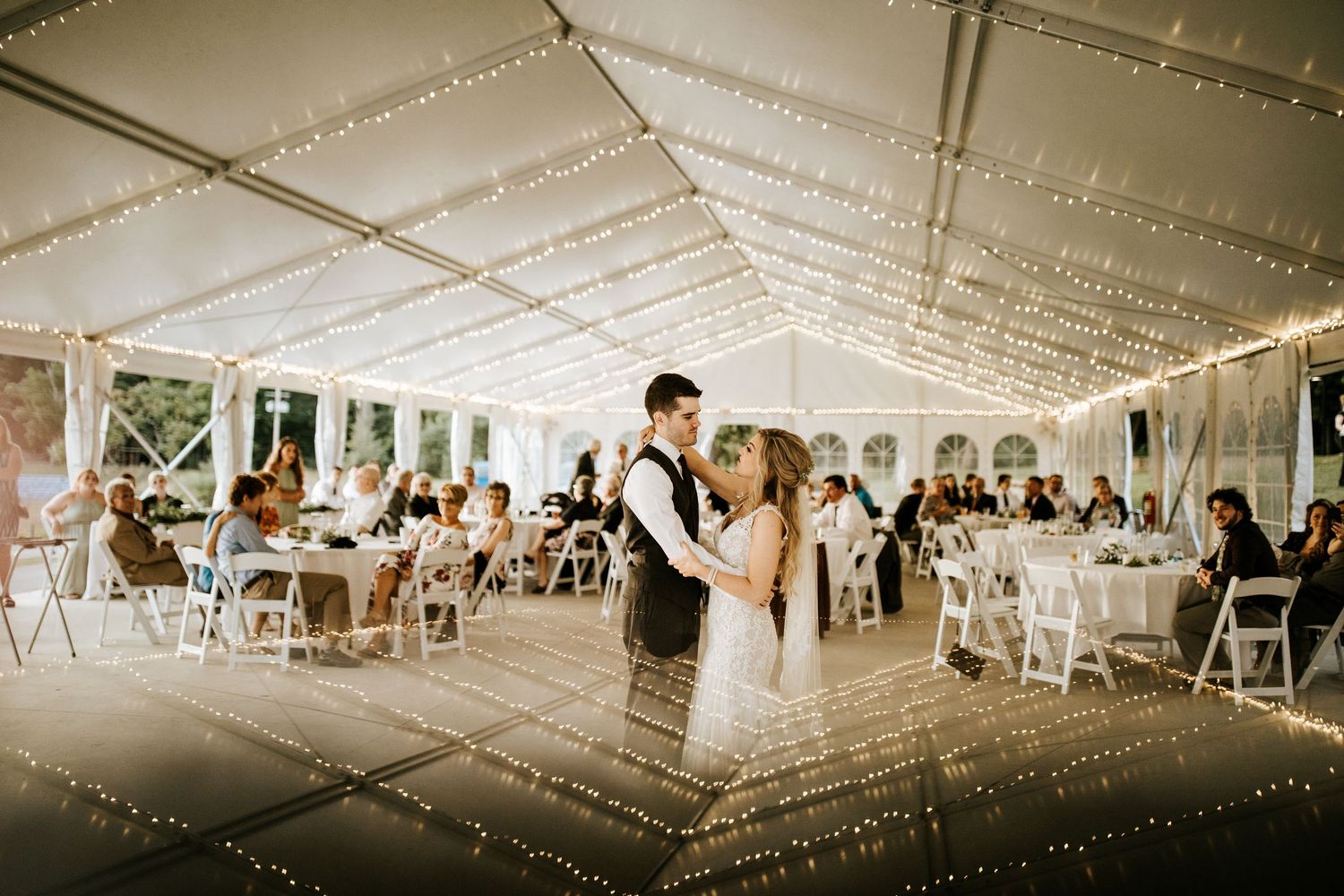 Wedding celebration in a spacious white tent decorated with romantic overhead lighting and white chairs.