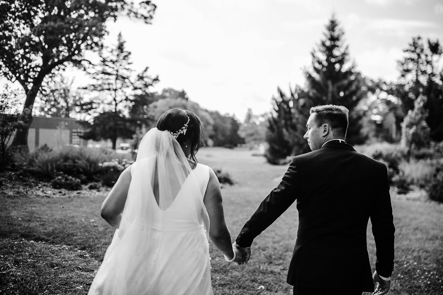 Black and white photo of newlyweds walking hand in hand on a garden path.