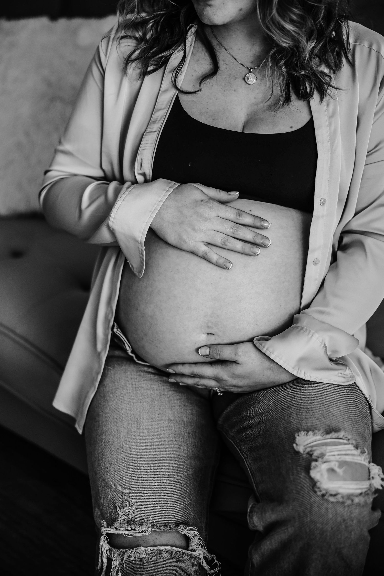 Black and white maternity photo showing hands cradling a pregnant belly while wearing ripped jeans and a blazer.