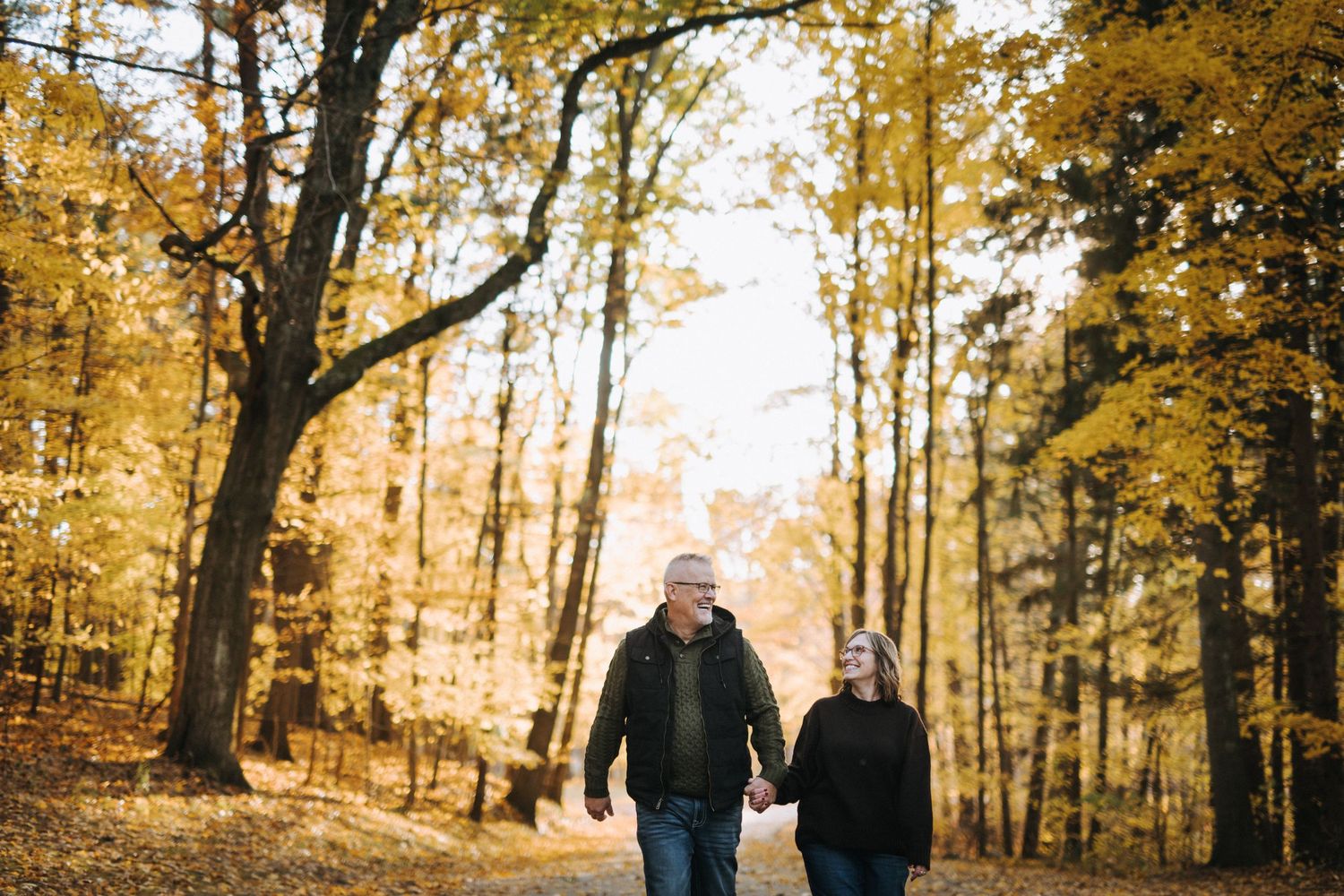A couple walks through a sunlit autumn forest with golden leaves and tall trees.