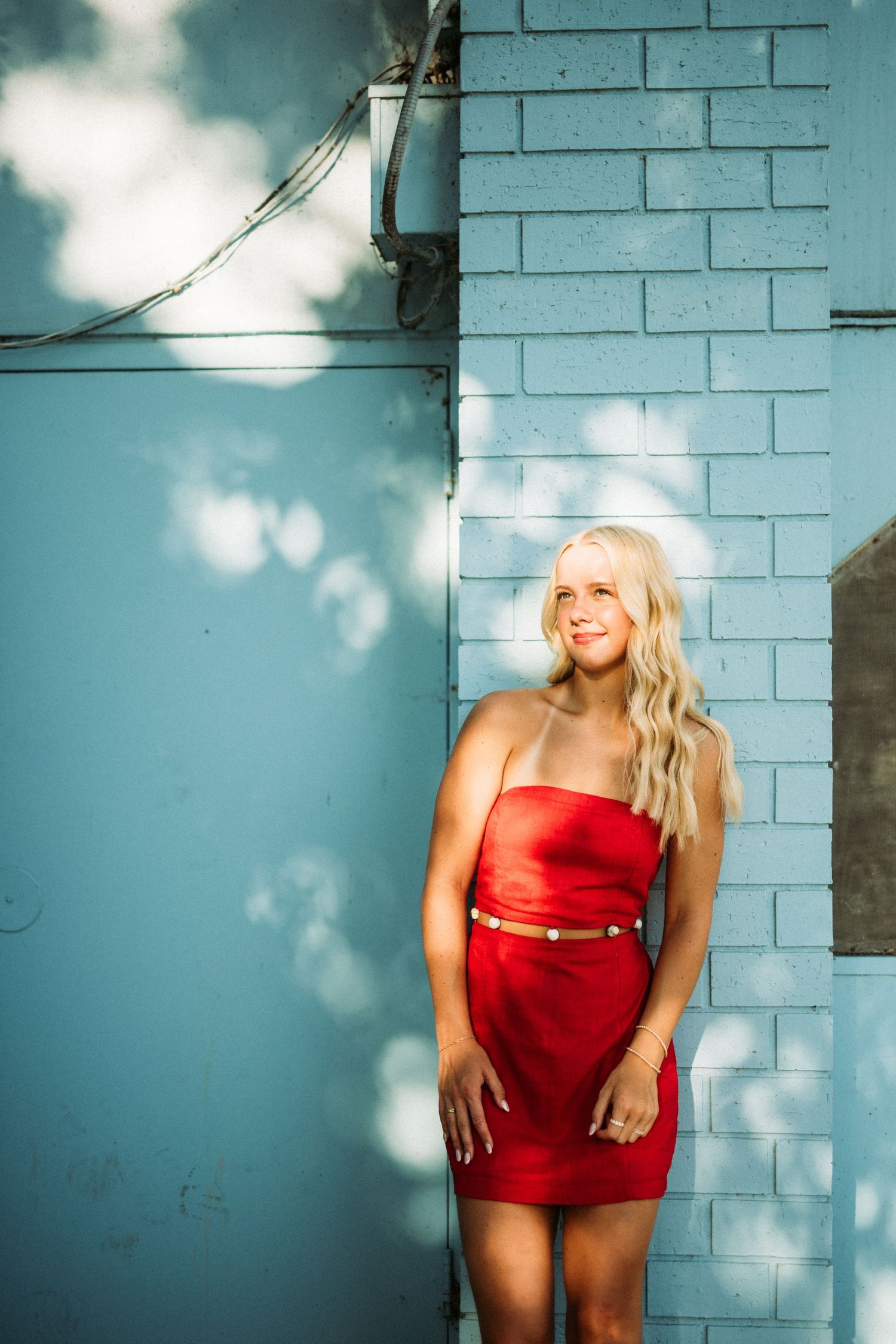 Woman in red dress poses against turquoise wall with dramatic shadows and lighting.