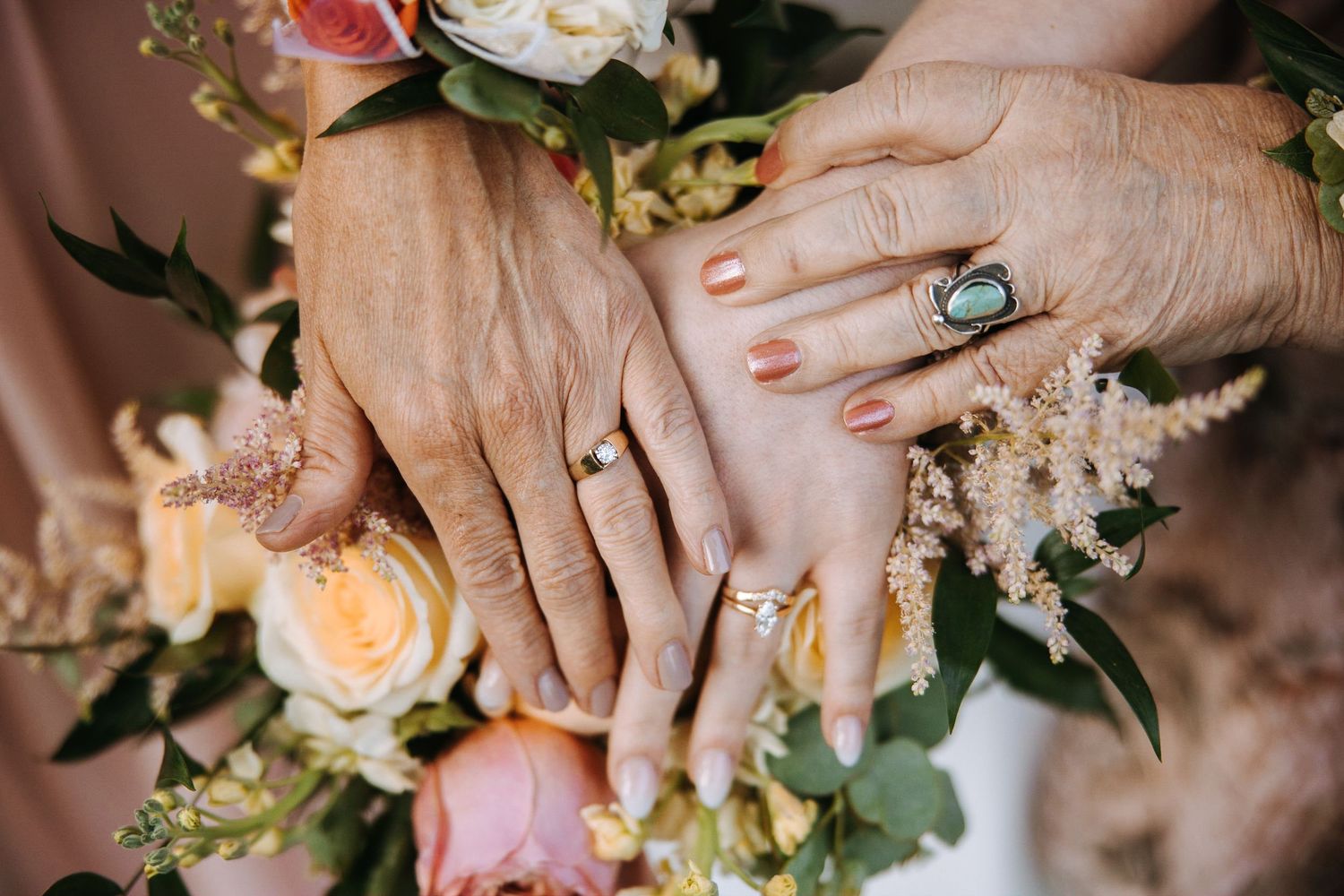 Close-up of hands with wedding rings resting on a bouquet of pink and cream roses.