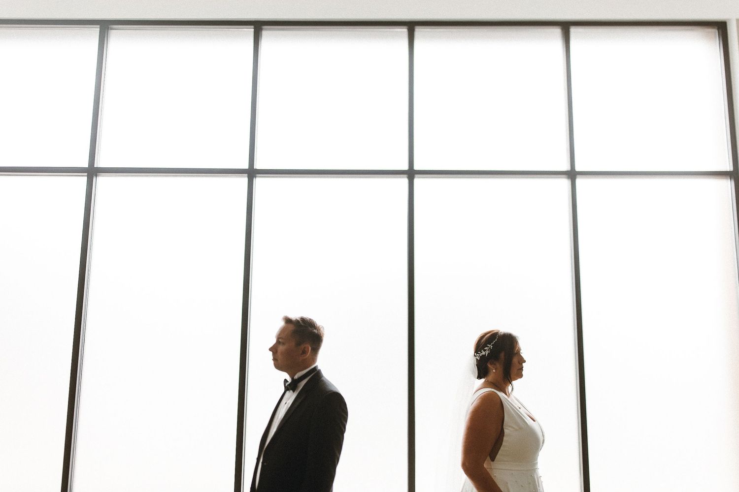 Dramatic silhouette of a couple standing back to back against large bright windows.