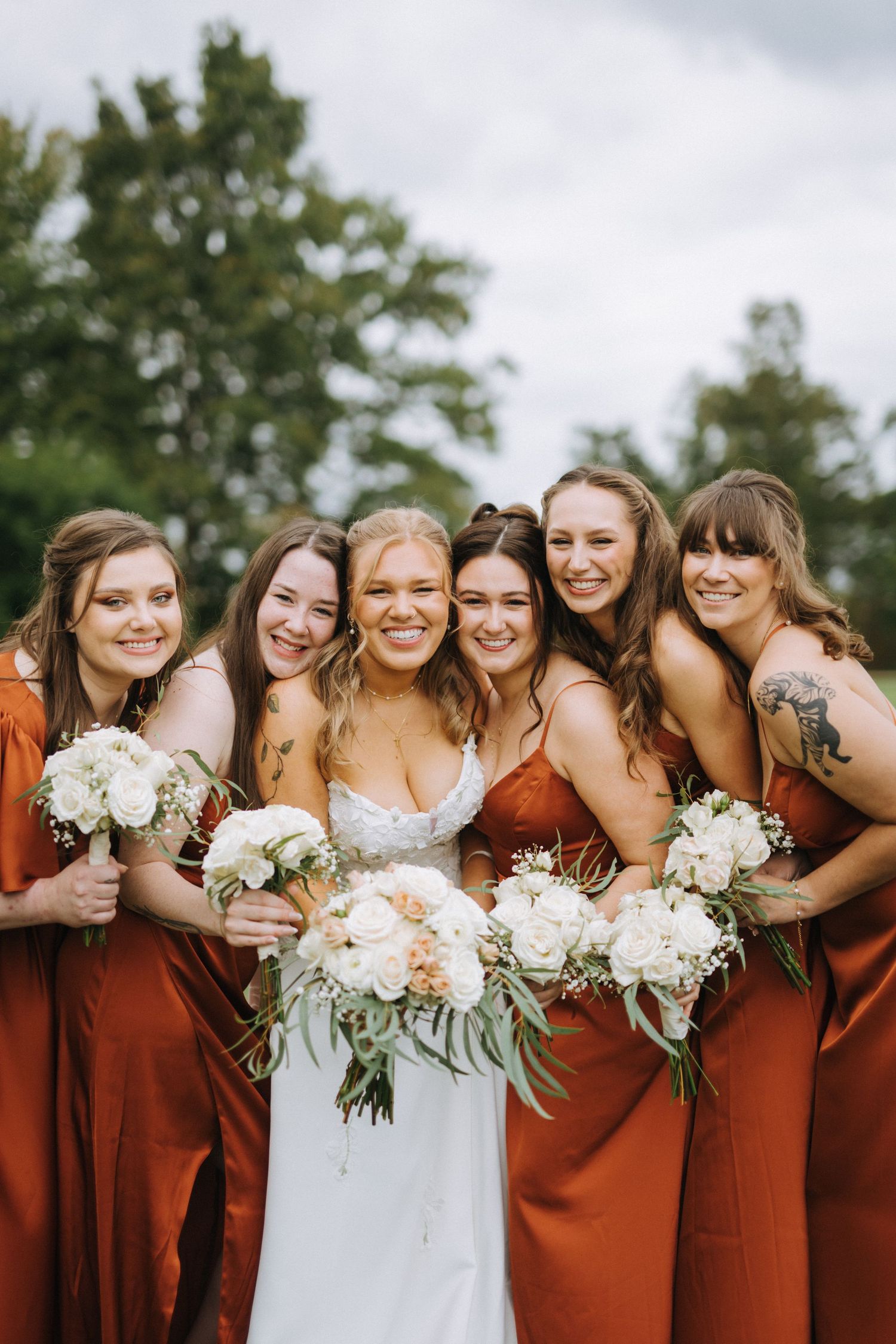 Bridesmaids in rust-colored dresses pose together holding white floral bouquets outdoors.