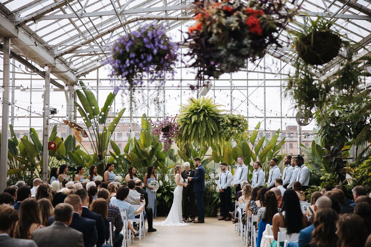 Wide angle view of a greenhouse wedding ceremony with guests seated and tropical plants overhead.