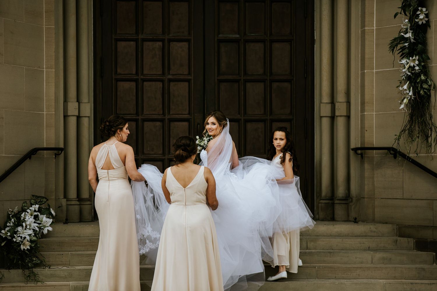 Bridesmaids in light colored dresses help arrange a wedding gown on church steps.