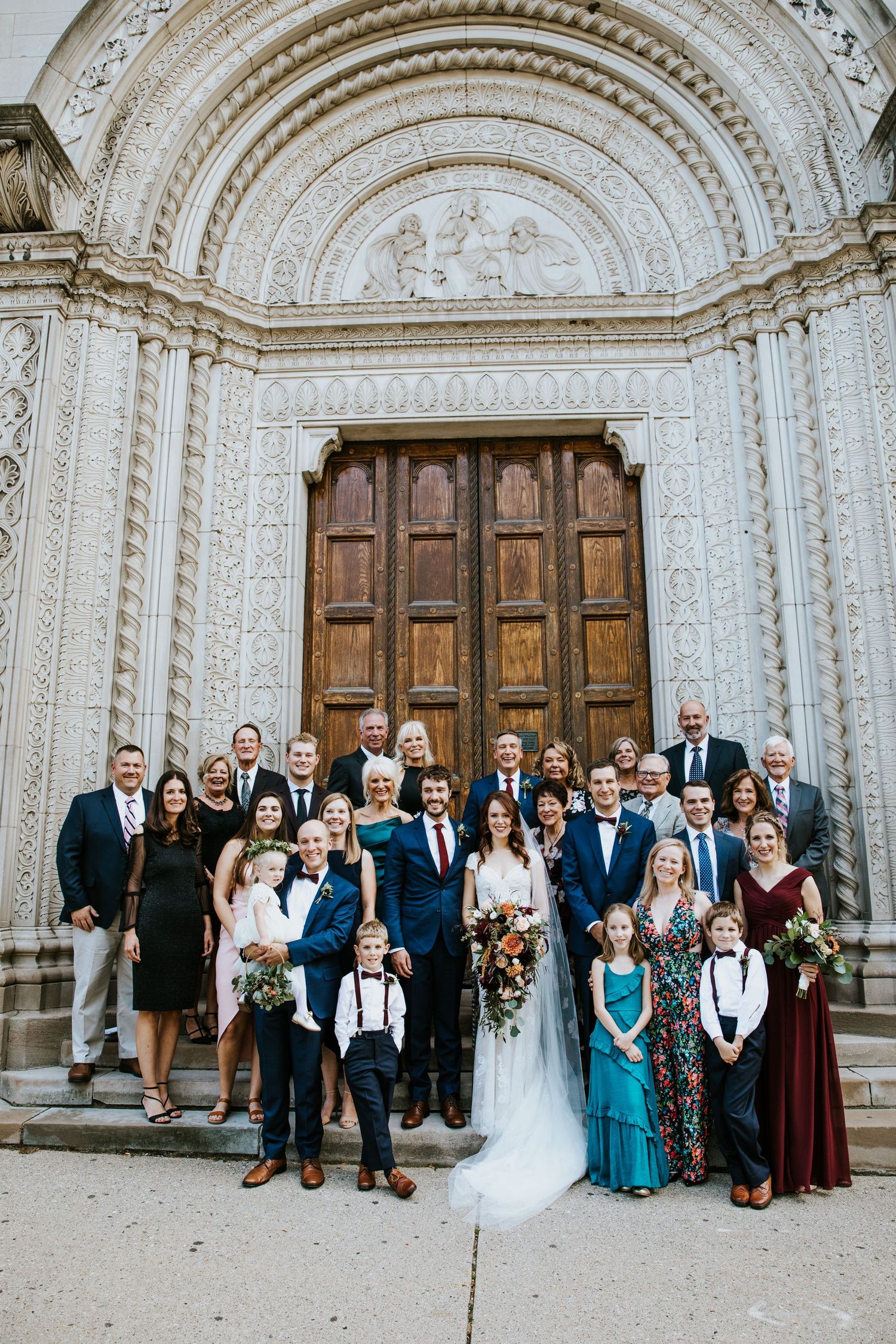 Wedding party gathered in front of ornate stone church with arched doorways.