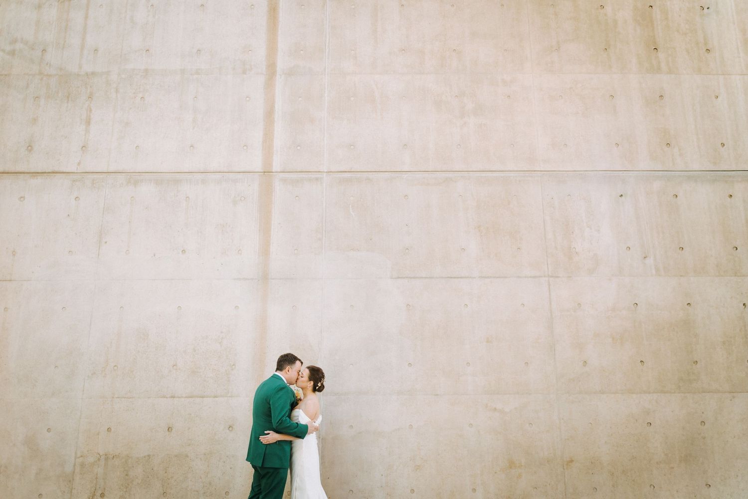 Bride and groom sharing a romantic kiss against a large concrete wall backdrop on their wedding day.