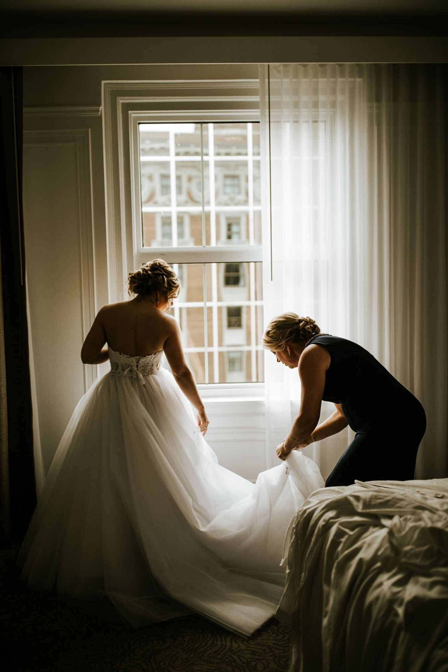 A wedding dress being adjusted in a sunlit room with dramatic window lighting.