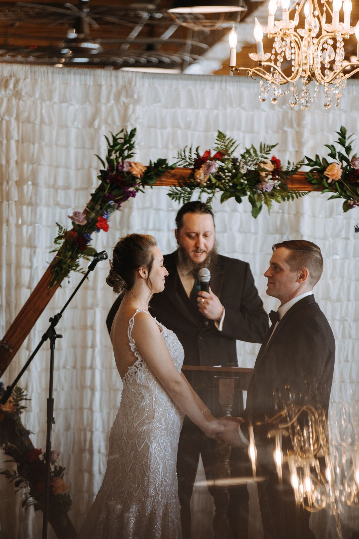 Wedding ceremony under a floral arch with elegant chandelier and white backdrop.