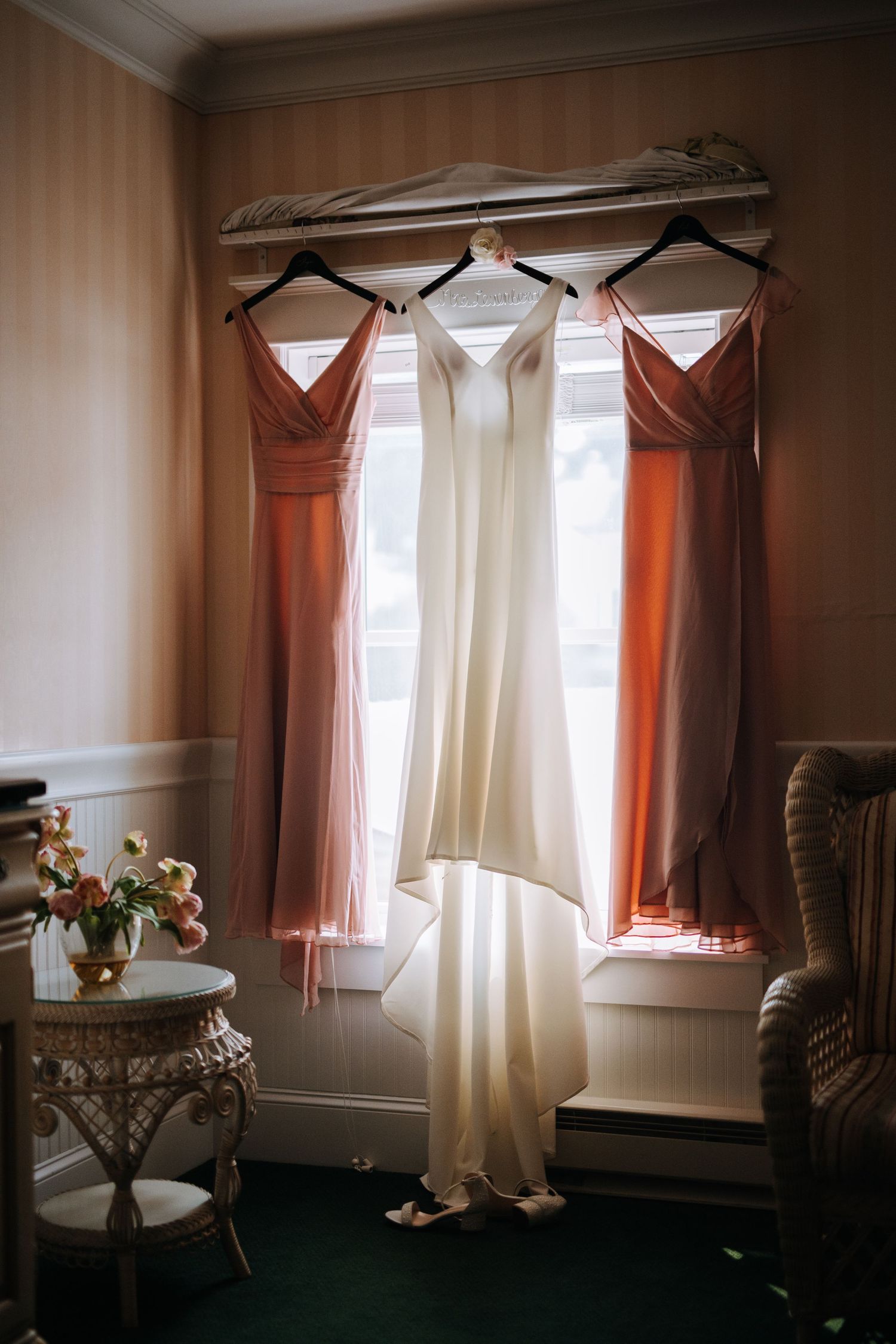 Wedding dress and two coral bridesmaid dresses hanging in a room with striped wallpaper.