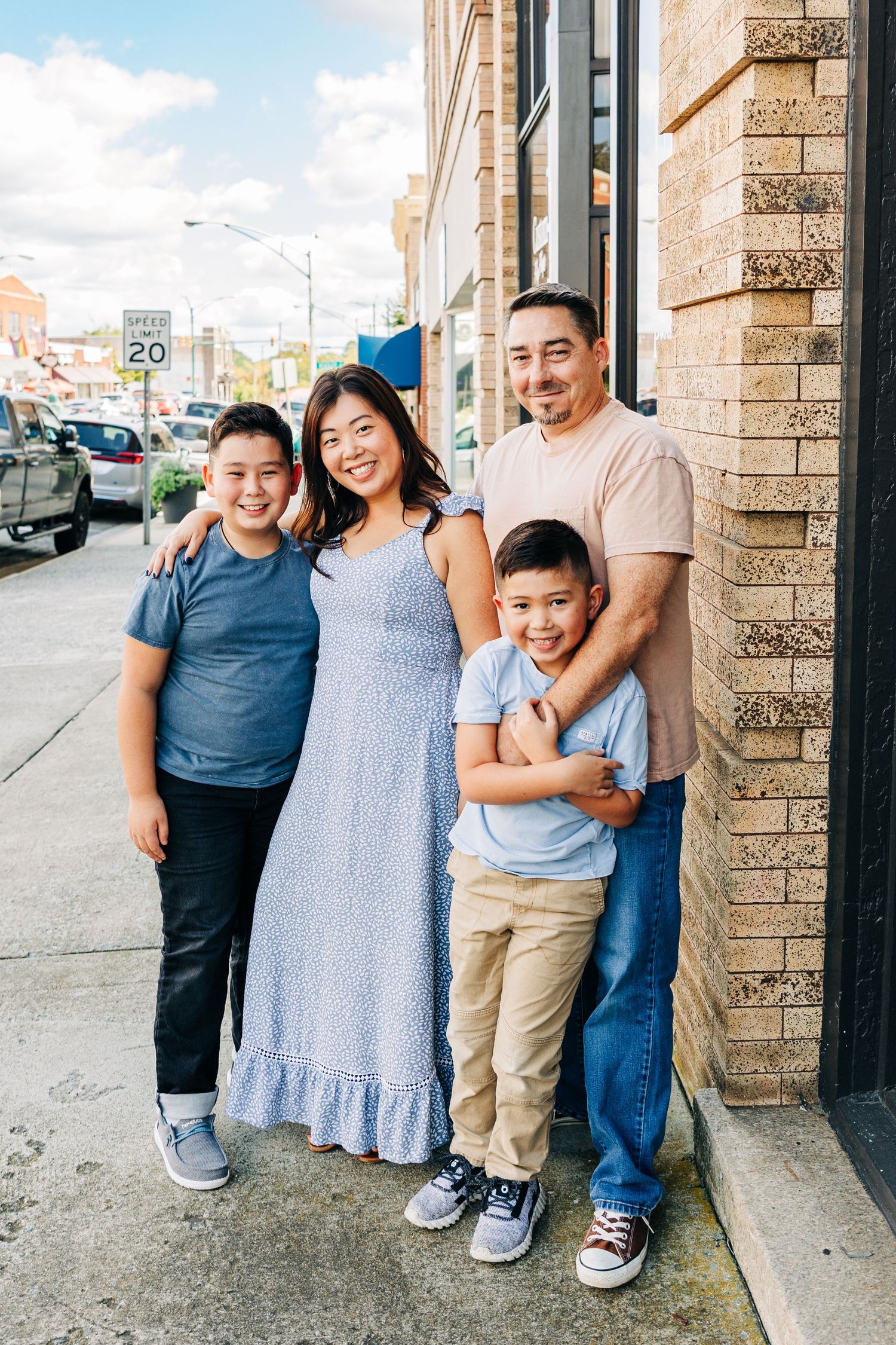 A family stands together smiling on a city sidewalk outside a brick building on a sunny day.