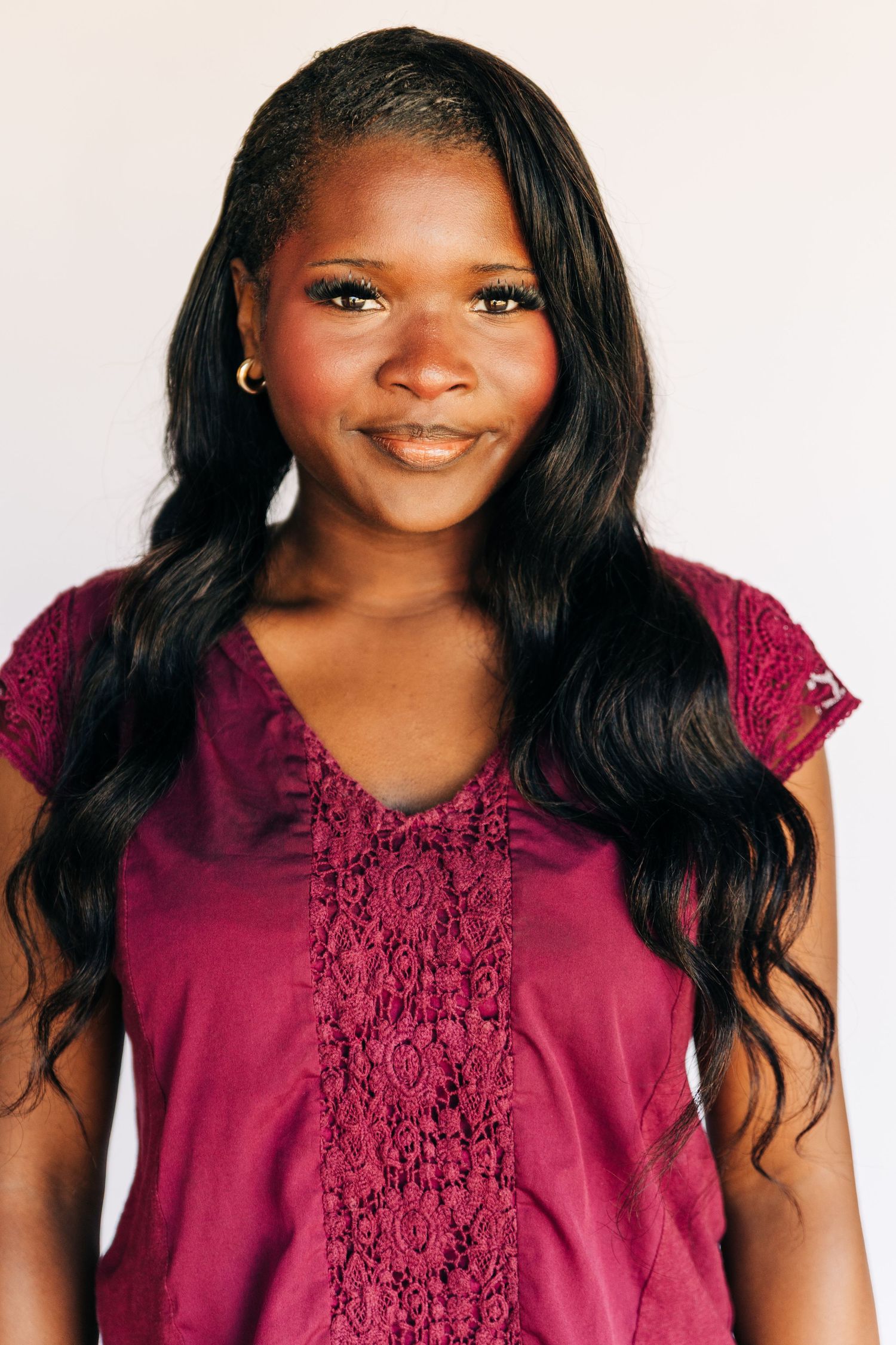Young woman with long wavy black hair smiling, wearing a burgundy lace top, against a light background.