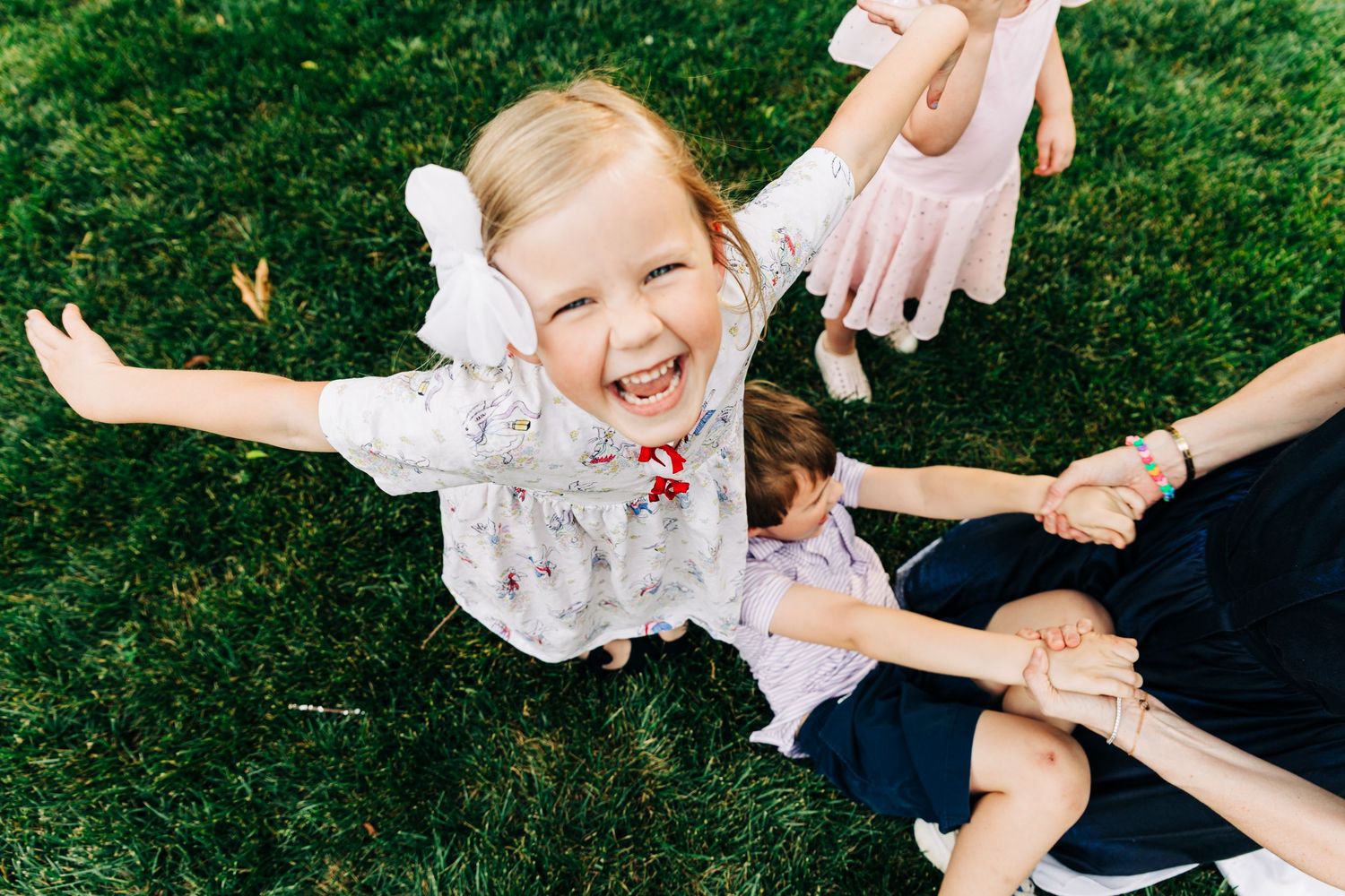 Joyful blonde toddler girl laughing with arms outstretched on green grass with family outdoors.