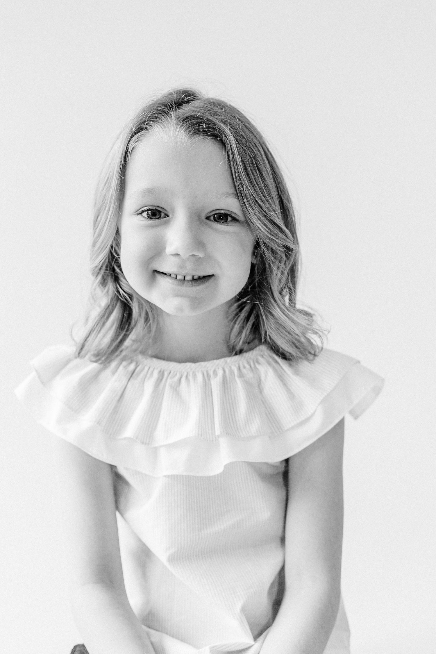 Black and white portrait of a child wearing a white ruffled top and smiling warmly at the camera.