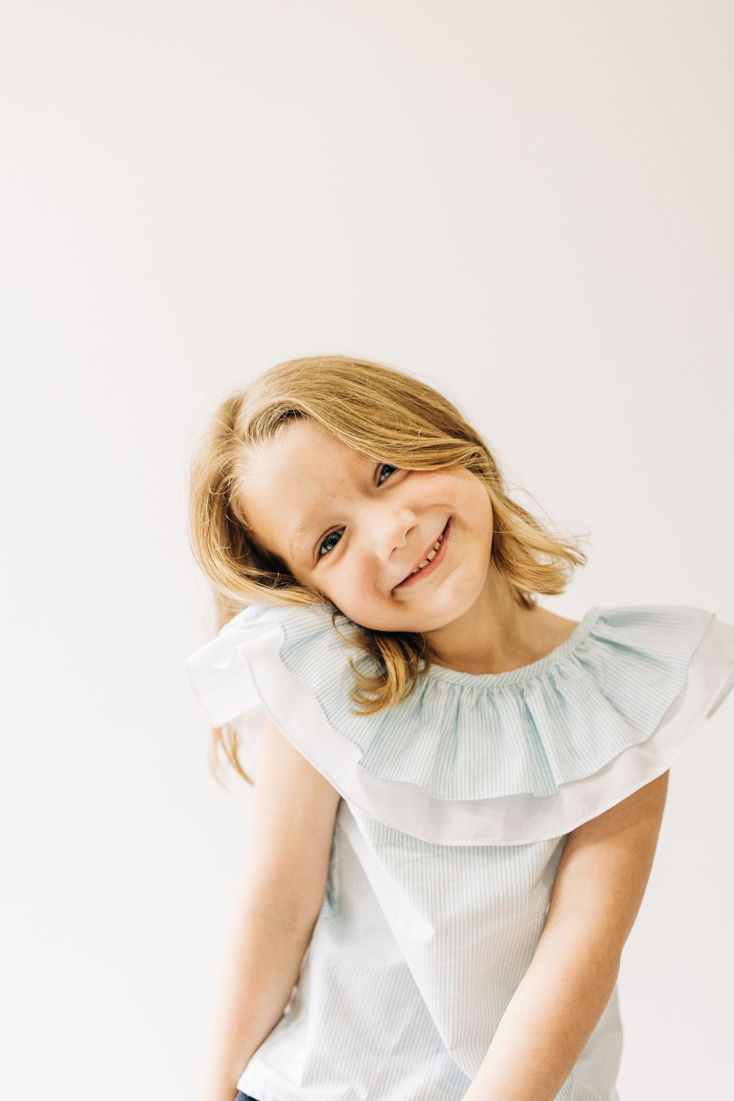 Photo of a young child in a white ruffled dress posing playfully against a light background.