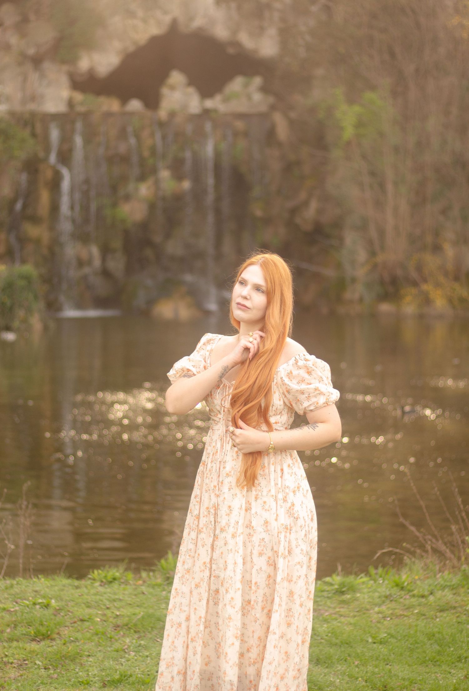 A woman with long red hair is standing by a calm body of water, wearing a floral dress. In the background, there is a waterfall.