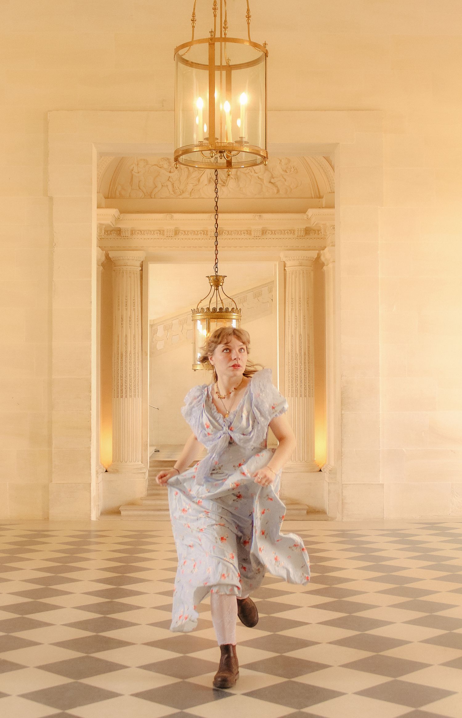 A woman in a floral dress is running in an ornate piece with chandeliers and light-colored walls.