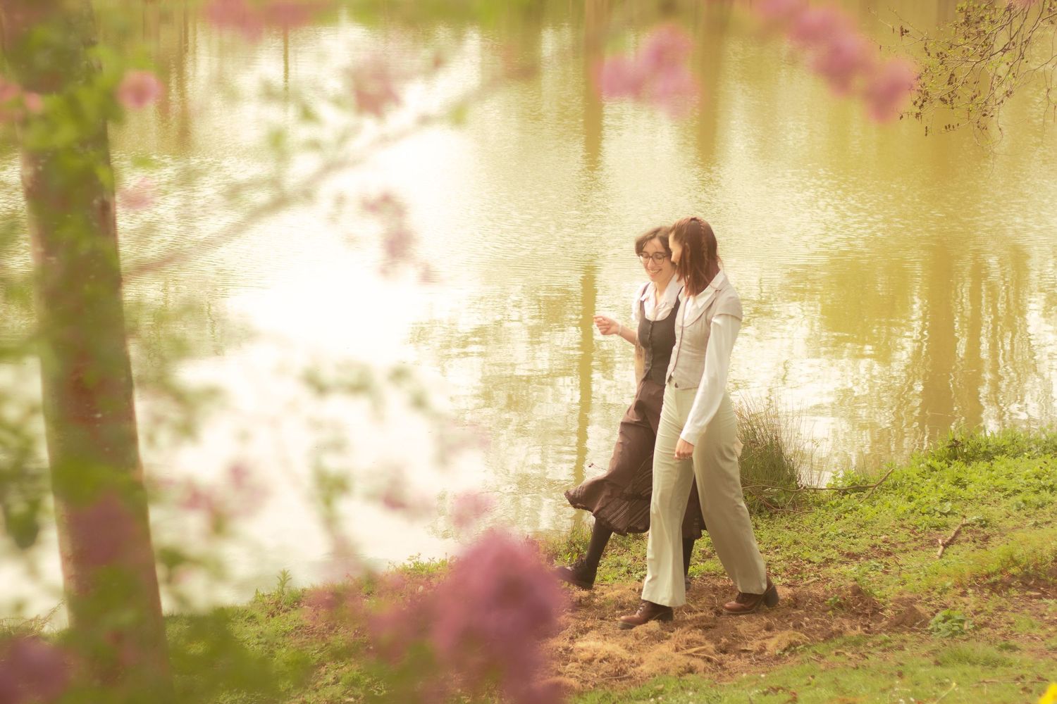 Two girls are walking and talking, in the background there is a calm body of water.
