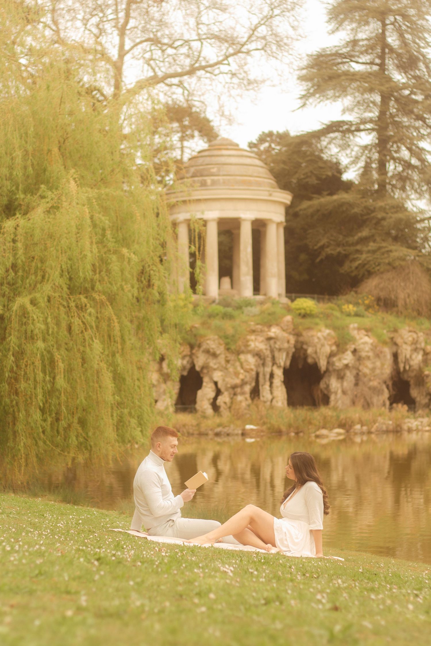 A couple is sitting in the grass reading a book, in the background there is a willow tree and a temple located in Bois de Vincennes.