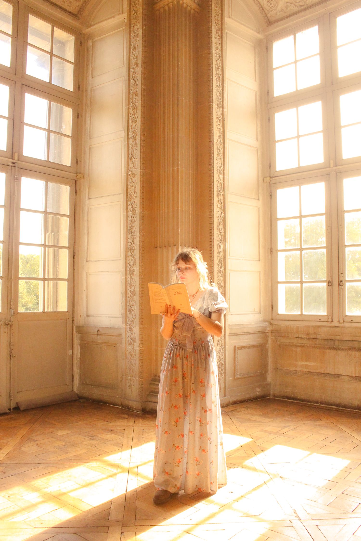 A girl is standing in a piece with long glass windows inside a baroque castle in Paris, she is reading a book and has a long dress.