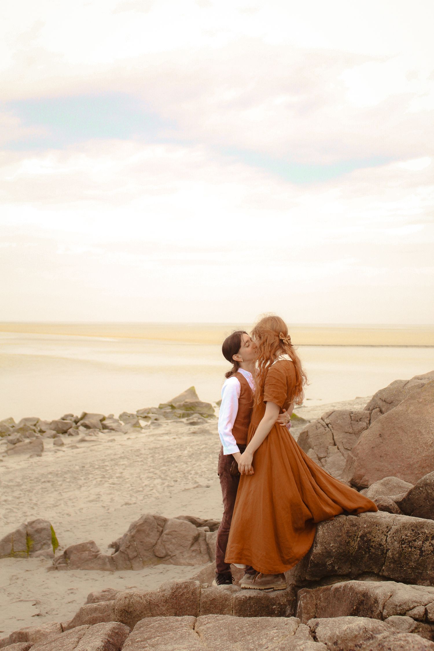 A couple is kissing, they are dressed in a vintage style, both are wearing brown. They are located in a beach, standing by the rocks.