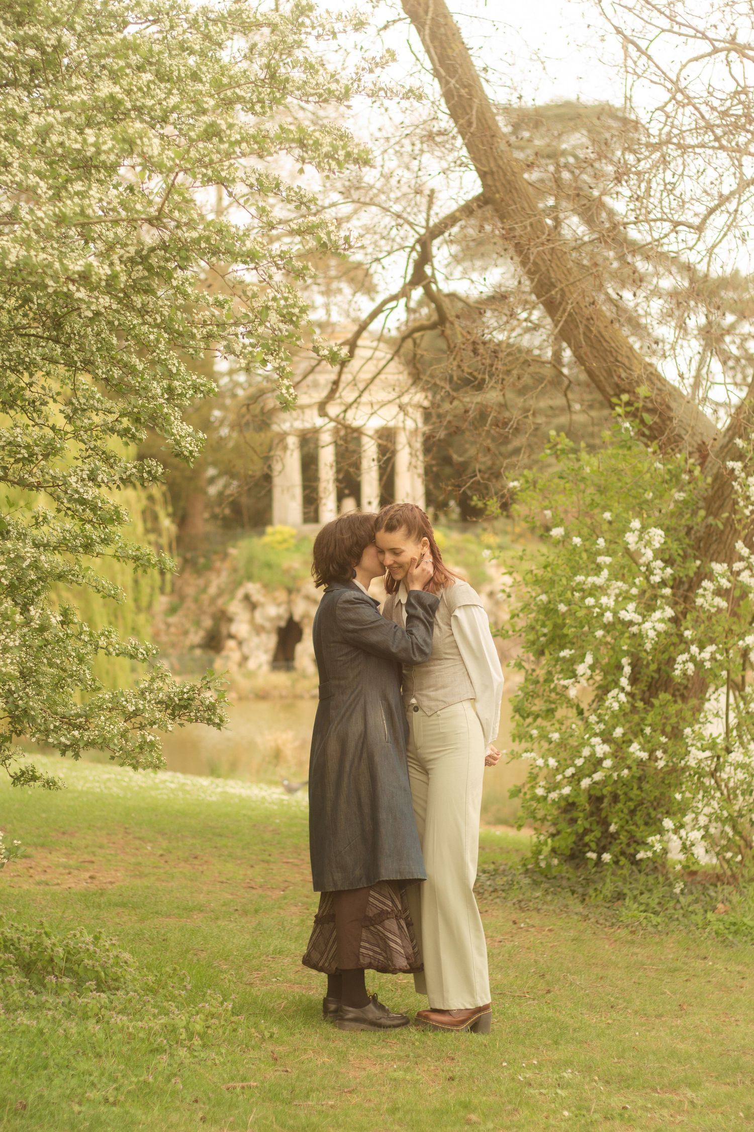 A lesbian couple are standing near to each other, one is whispering in the other's ear. In the background there is a temple.