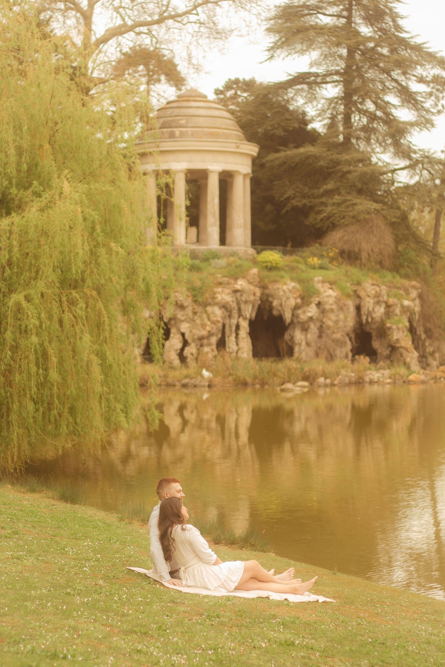 A couple is sitting in the grass, in the background there is a willow tree and a temple located in Bois de Vincennes.