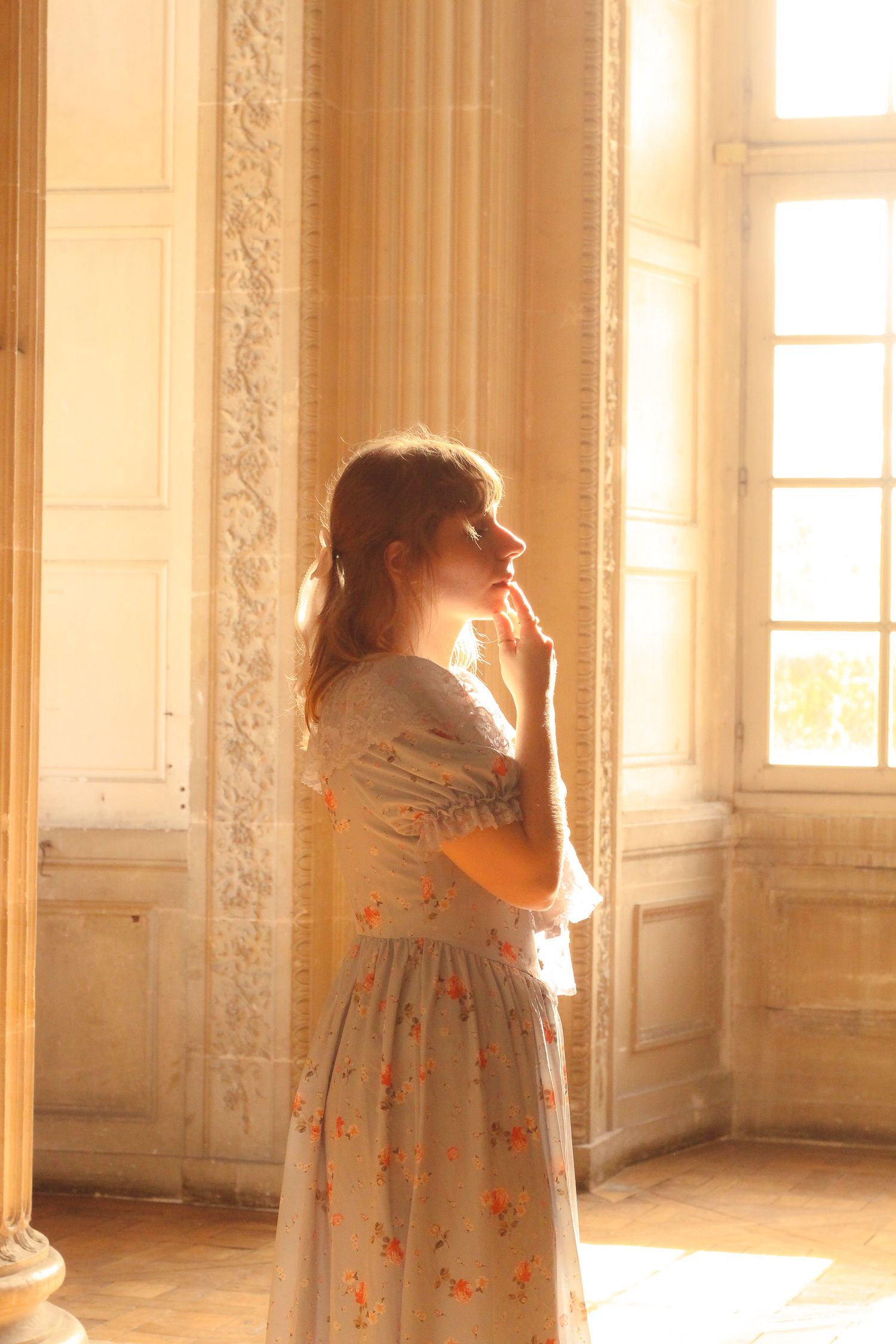 A girl is standing in a piece with long glass windows inside a baroque castle in Paris, she is blonde and has a long dress.