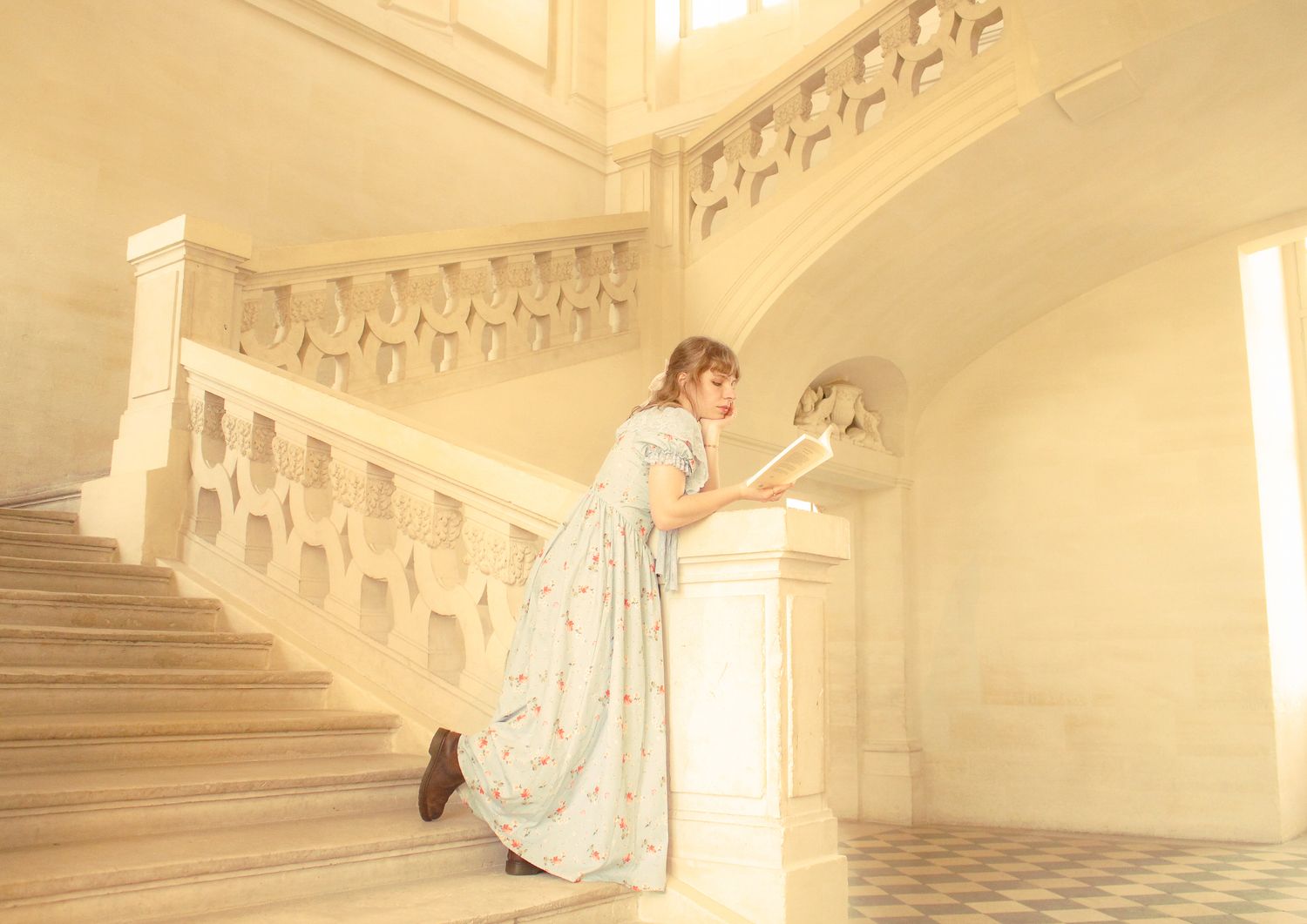 A woman in a floral dress leans against a white railing while reading a book on an ornate staircase with light-colored walls.