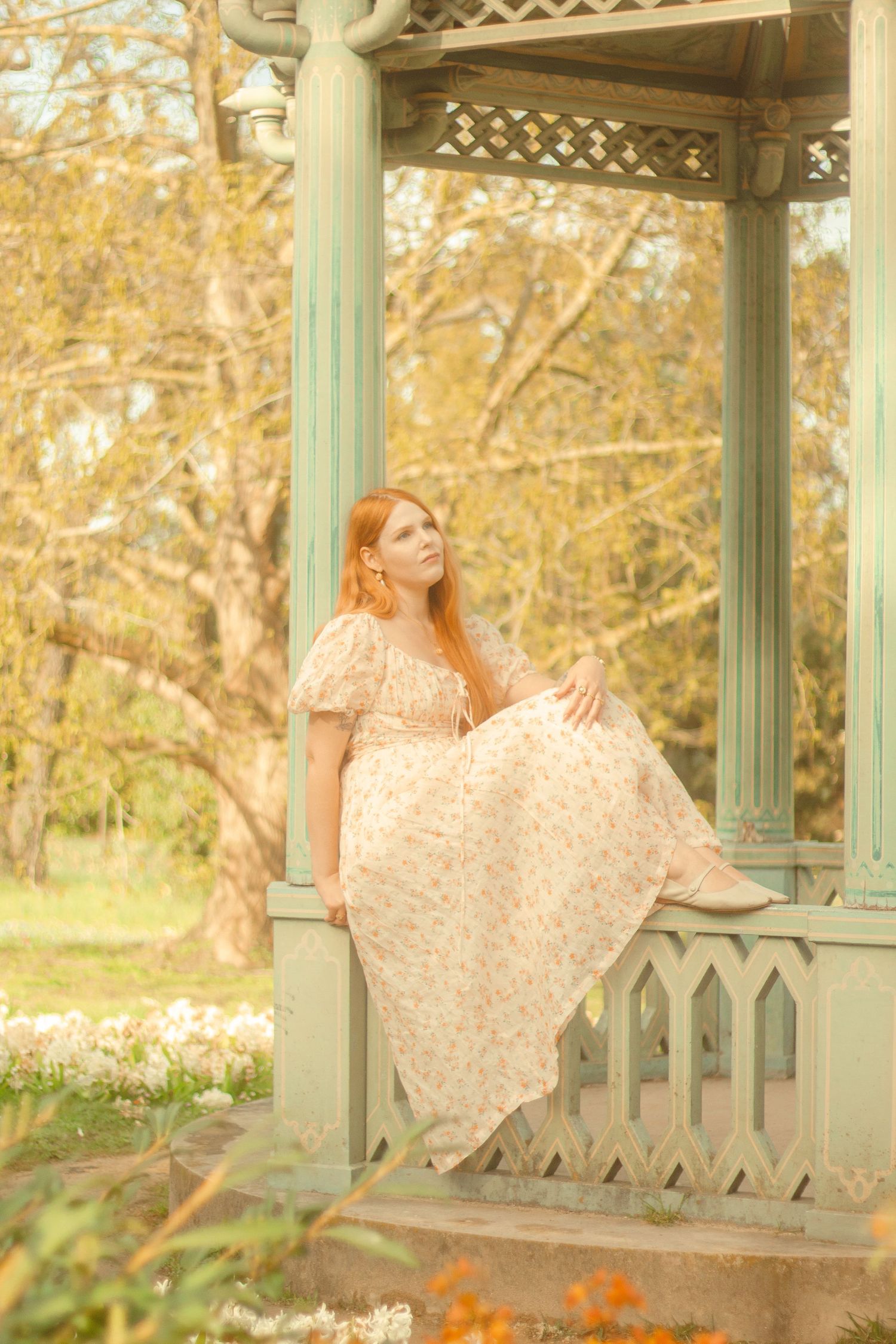 A woman with long red hair is sitting in a decorative gazebo, wearing a floral dress. She is surrounded by a garden filled with flowers.