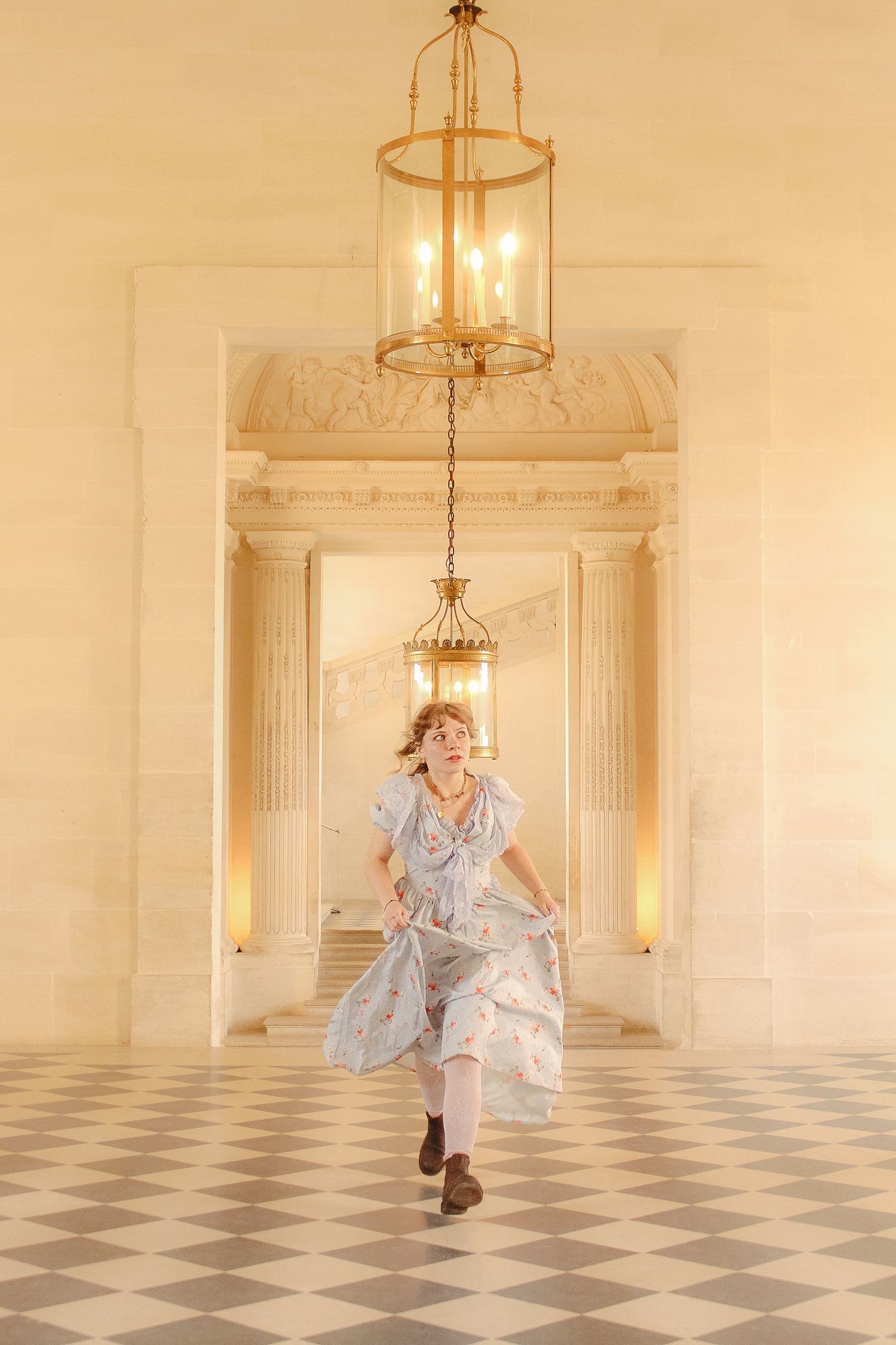 A woman in a floral dress is running in an ornate piece with chandeliers and light-colored walls.