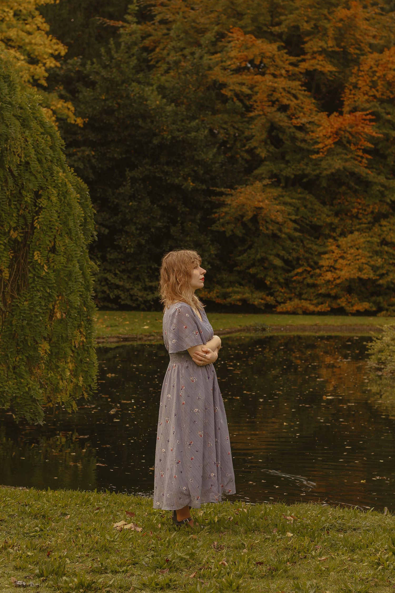A woman with short blonde hair is standing by a calm body of water, wearing a floral dress. The background colours is automnal.