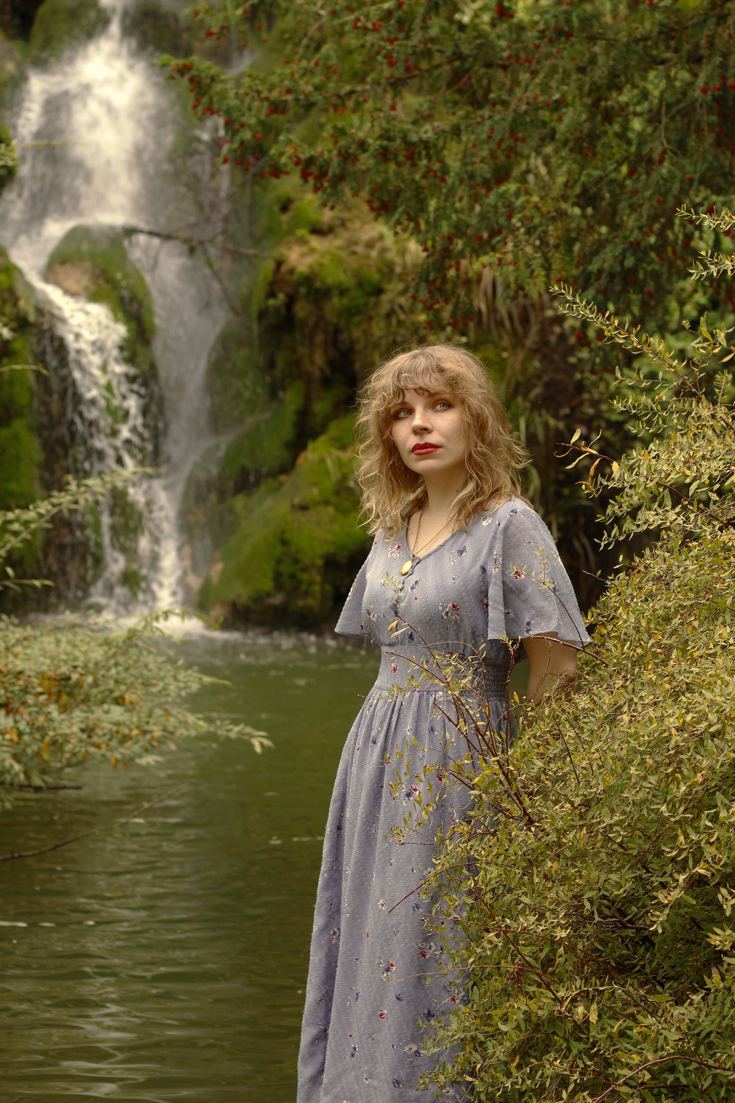 A woman with short blonde hair is standing by a calm body of water, wearing a floral dress. There is a waterfall in the background.
