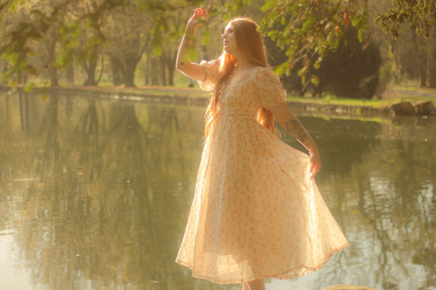 A woman with long red hair is standing by a calm body of water, wearing a floral dress.