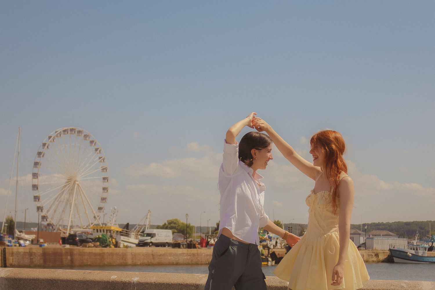 A partner makes their girlfriend twirl, she has a short   yellow dressed, in the background there is a ferris wheel and water
